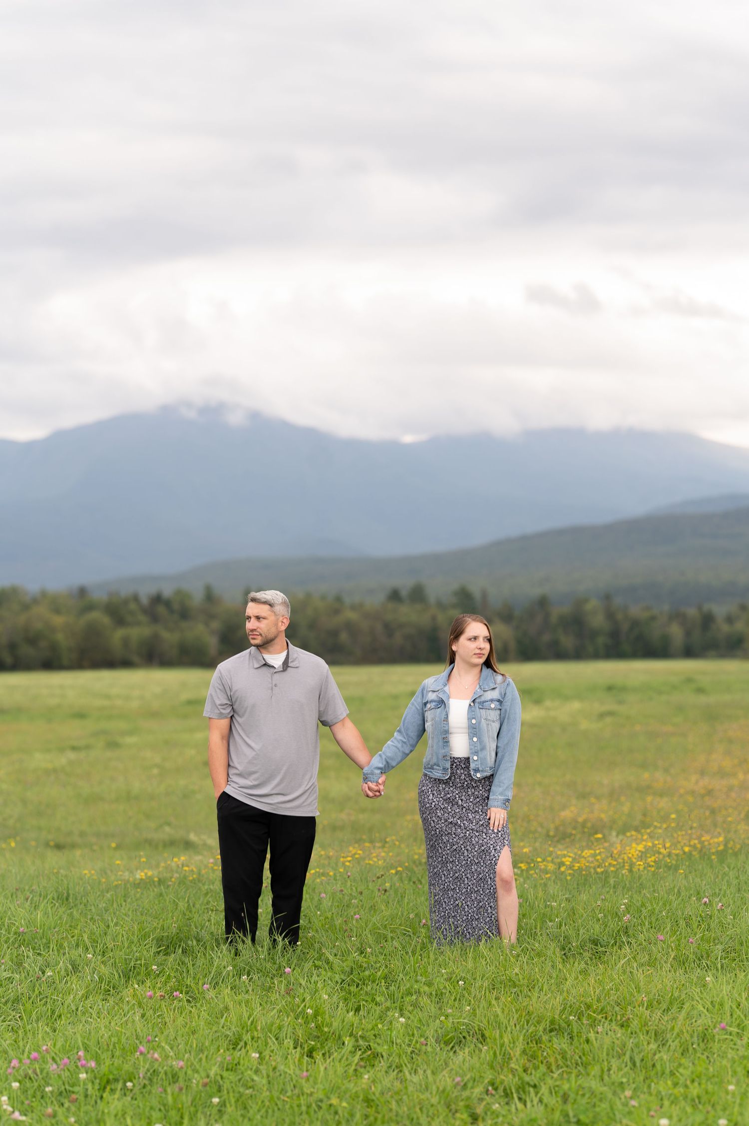Couple holding hands walking through green meadow with dramatic mountain scenery.
