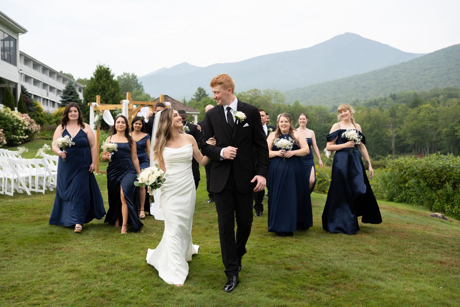Bride and groom walking with bridesmaids in navy dresses with mountain backdrop.