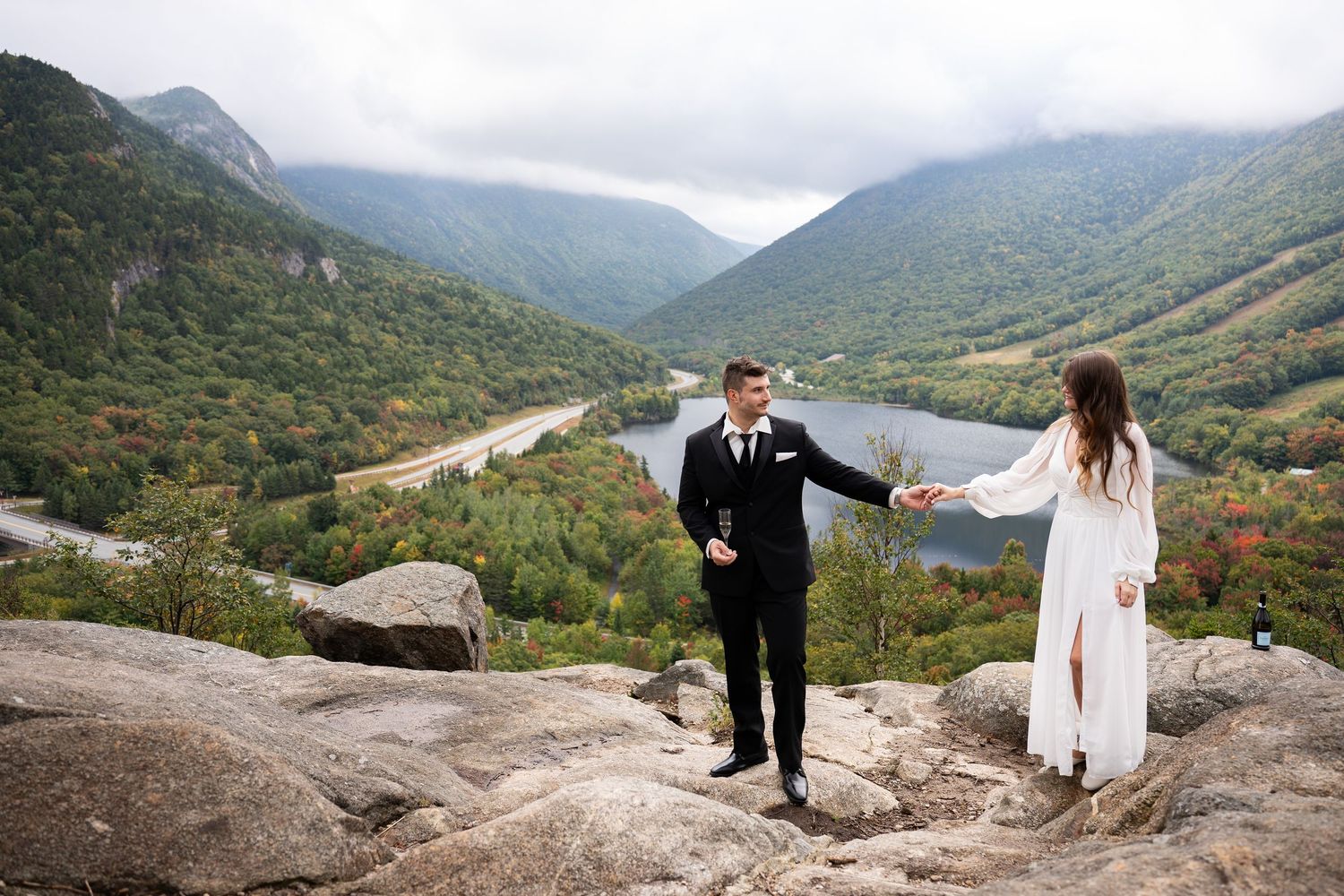 A bride and groom hold hands on a rocky mountain overlook with a scenic valley and river below on a cloudy day.