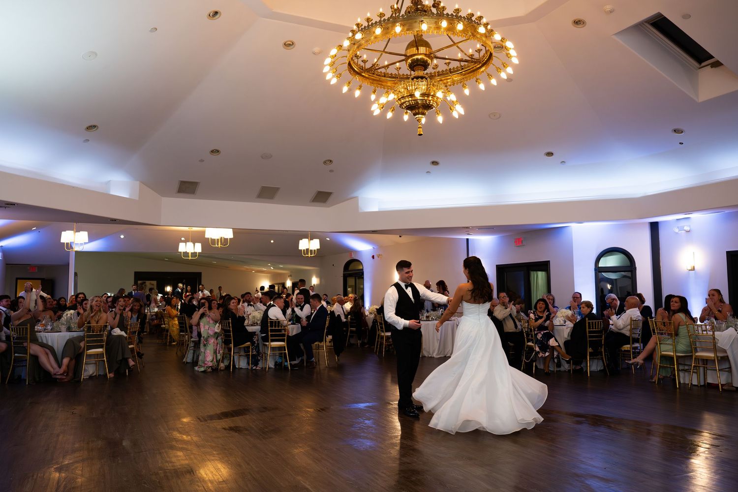 Bride and groom share their first dance under a golden chandelier in an elegant ballroom filled with wedding guests.