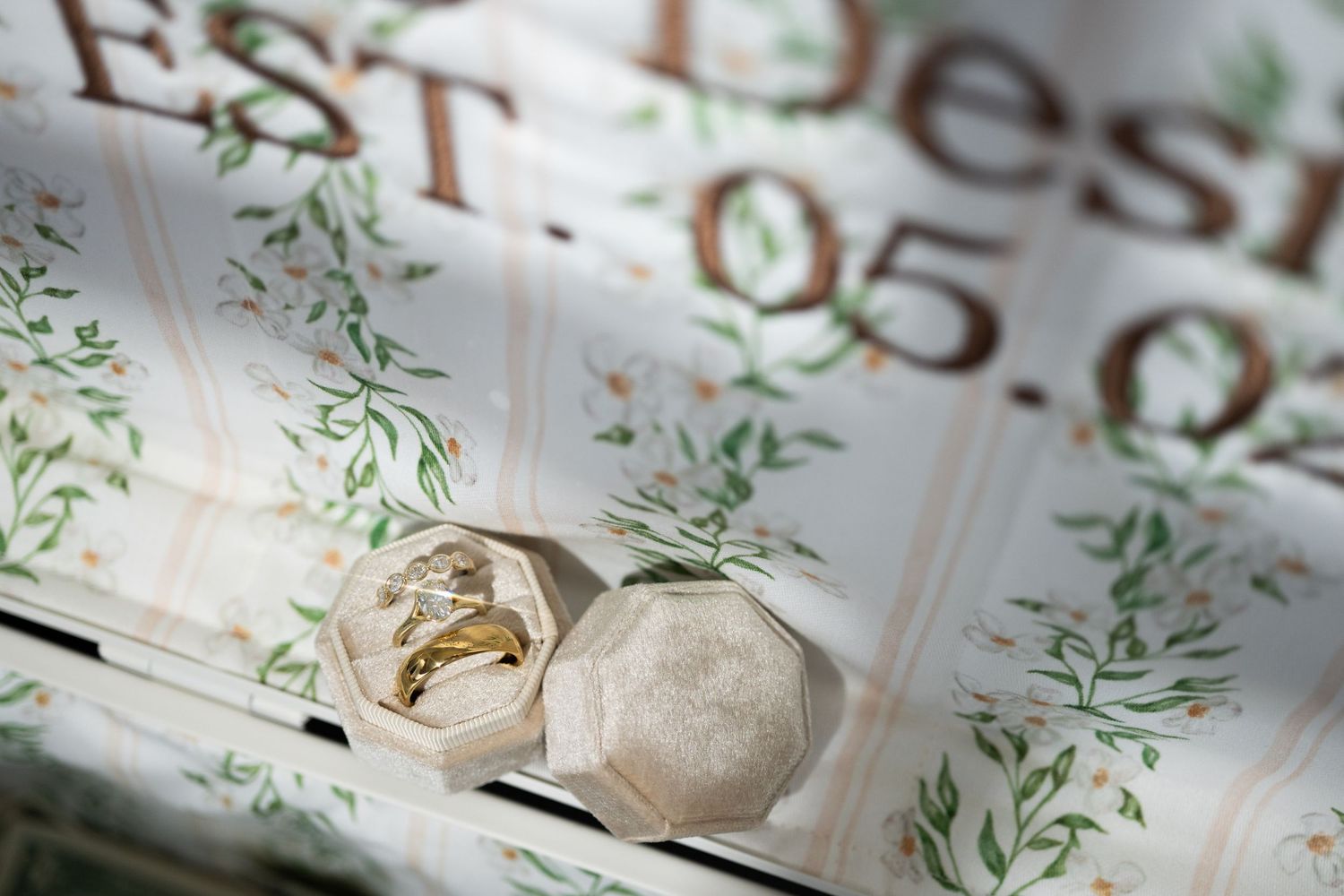 Gold wedding rings displayed in a velvet hexagonal box atop floral wedding stationery with green leaf details.