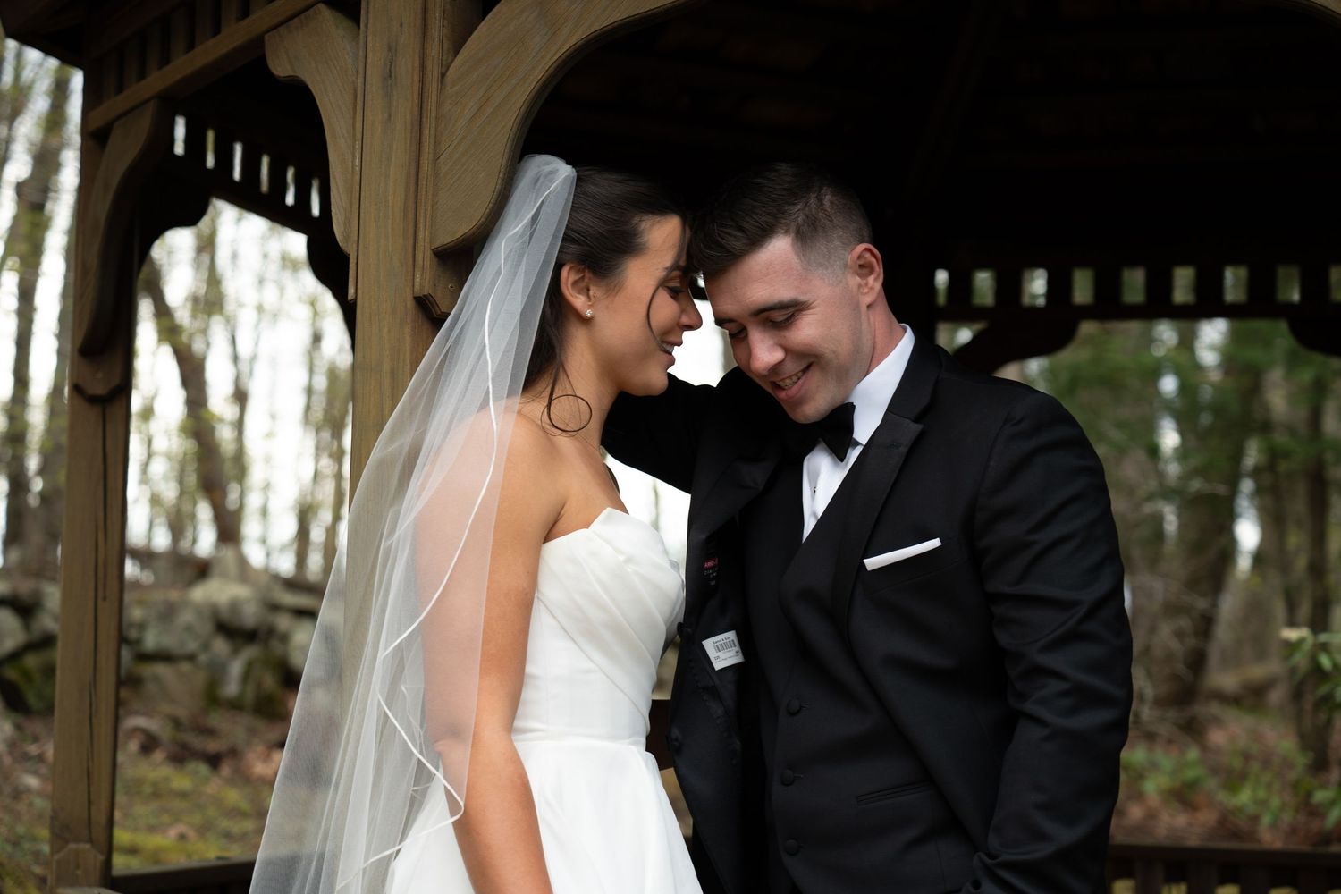 Happy bride and groom touching foreheads and smiling under wooden gazebo in wooded outdoor setting.