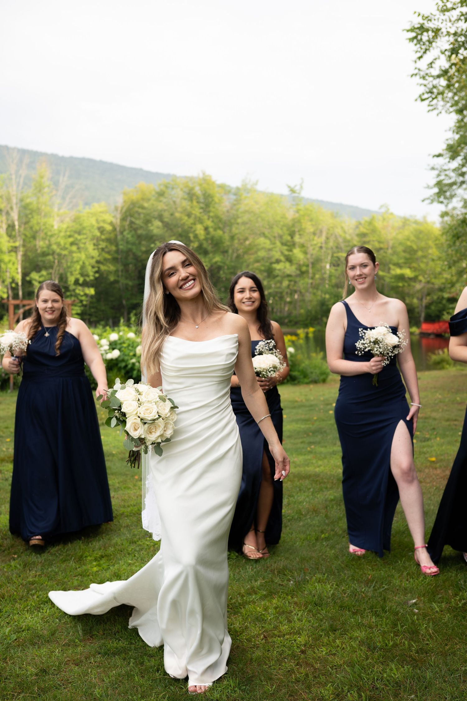 Smiling bride in white gown leading bridesmaids in navy dresses across green lawn.