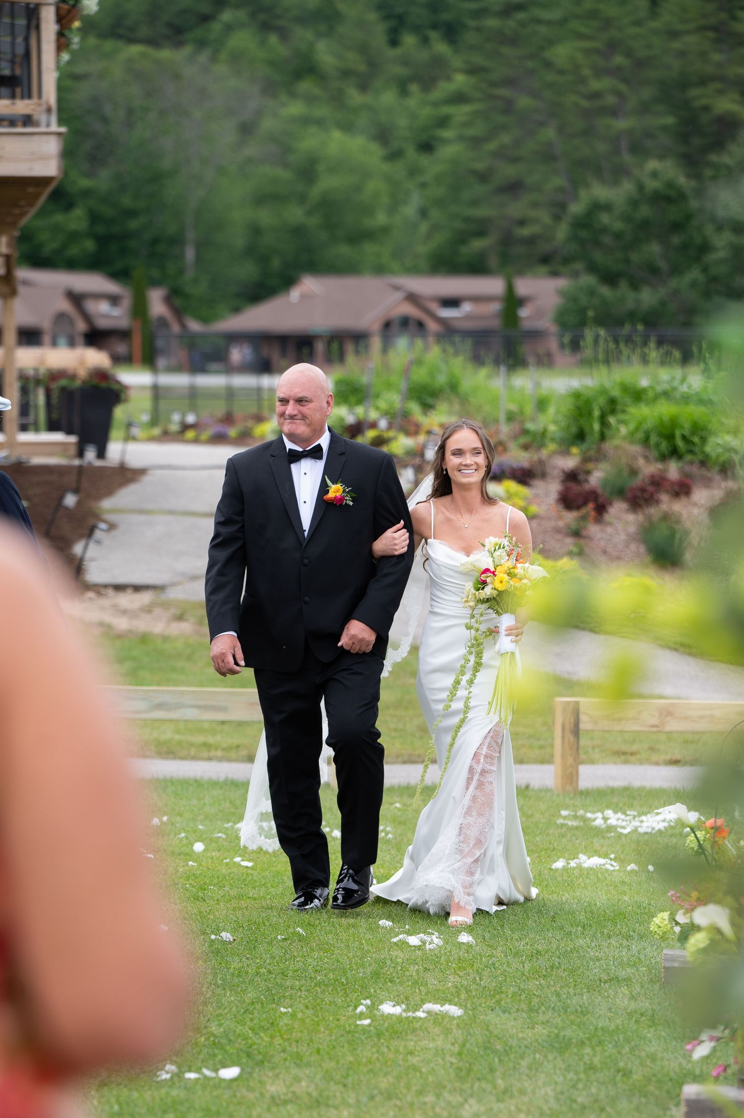 Bride in white gown walks down outdoor aisle with escort at elegant garden wedding ceremony.