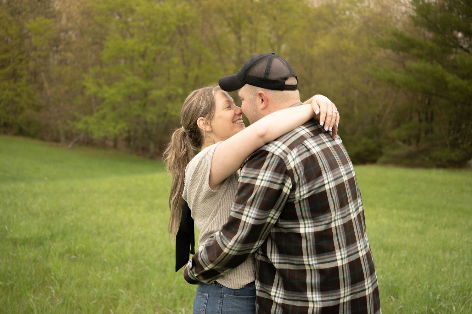 A smiling couple embraces in a green field surrounded by trees.