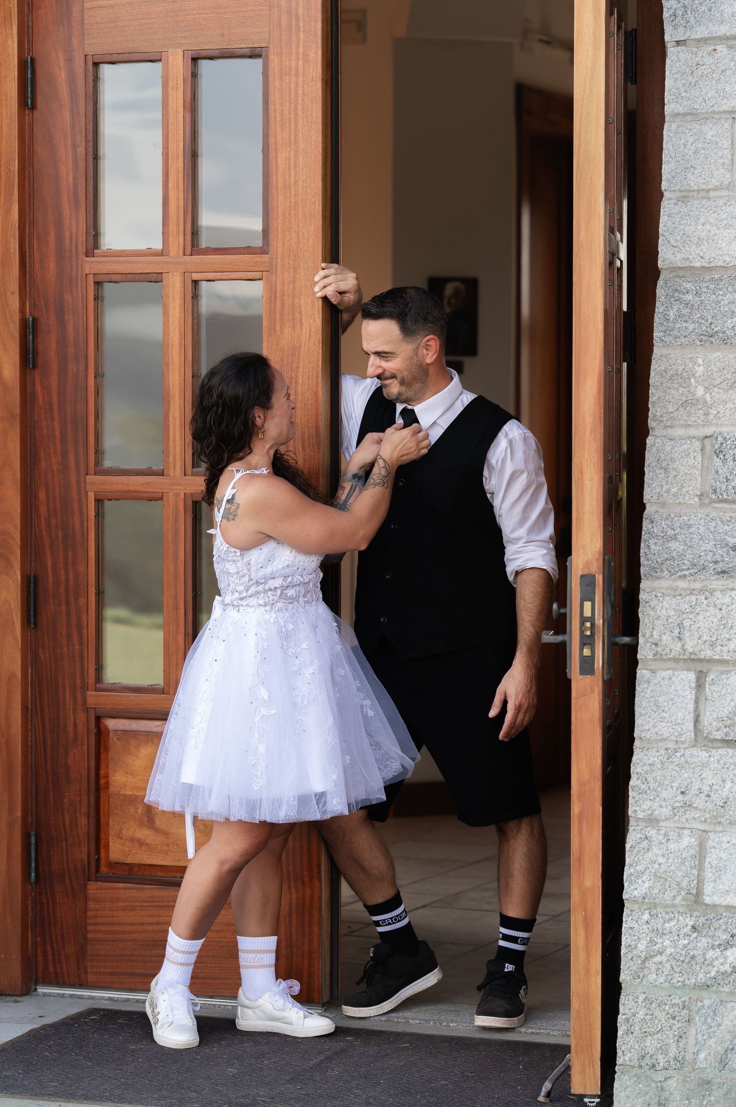 Bride in short white dress adjusts grooms tie as they stand together in a wooden chapel doorway.