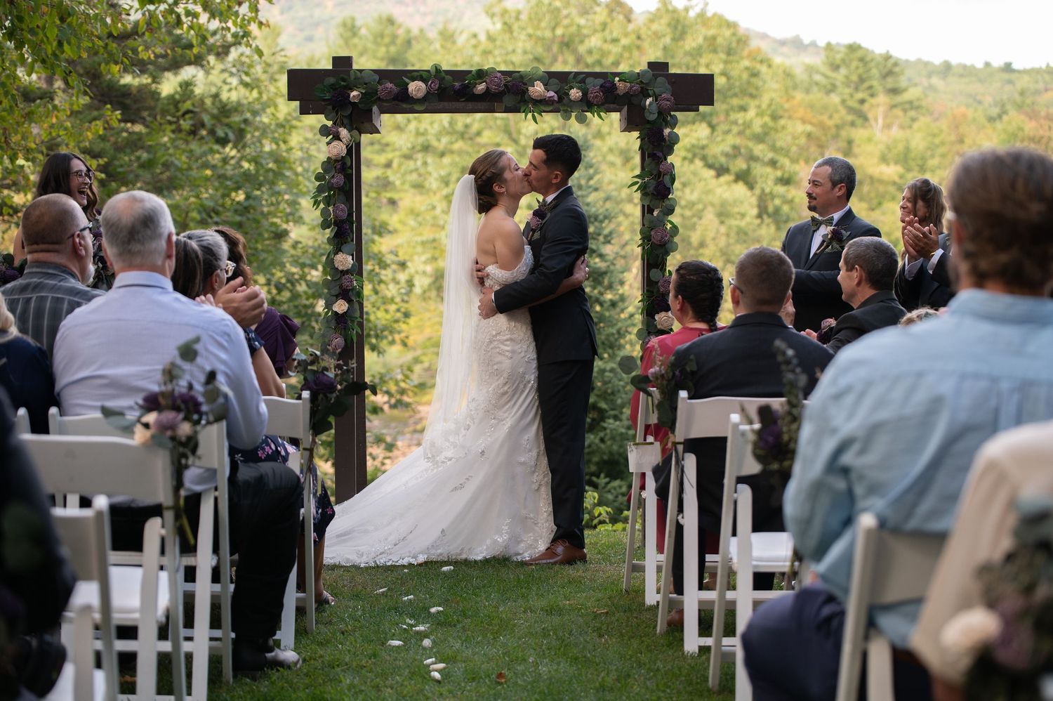 Bride and groom share first kiss under floral arch during outdoor wedding ceremony with seated guests.