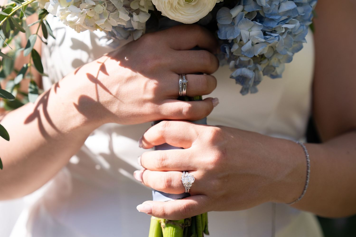Two brides holding a blue and white wedding bouquet, showcasing diamond engagement rings and wedding bands.
