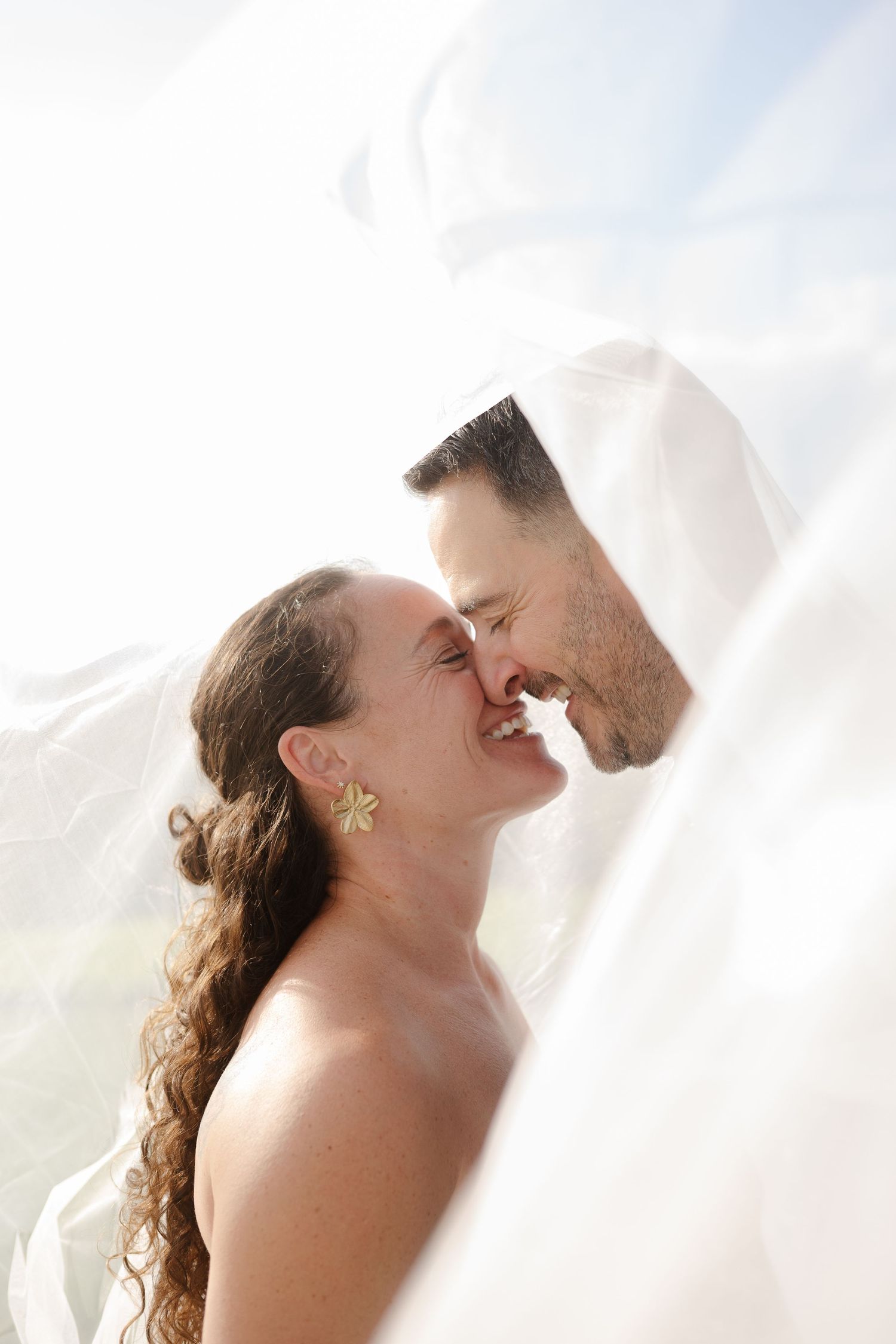 Bride and groom sharing a romantic kiss beneath a flowing wedding veil outdoors.