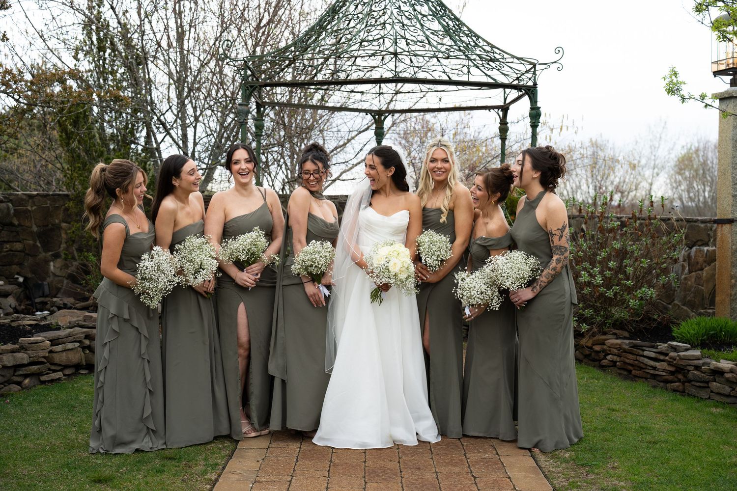 Bride in white gown standing with seven bridesmaids in sage green dresses holding white bouquets outdoors.