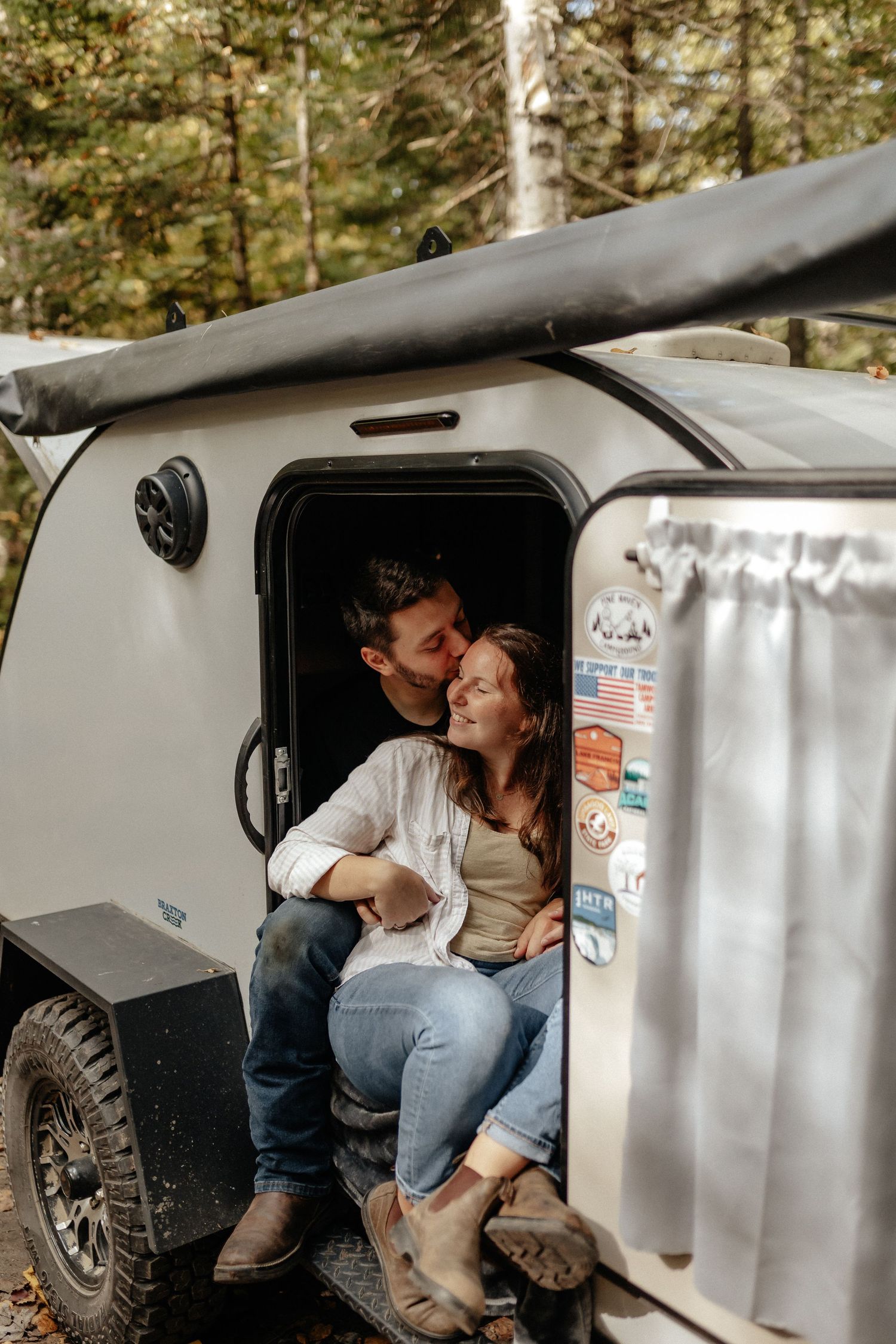 Couple sharing a tender kiss while sitting in the doorway of a teardrop camper trailer in a forest setting.