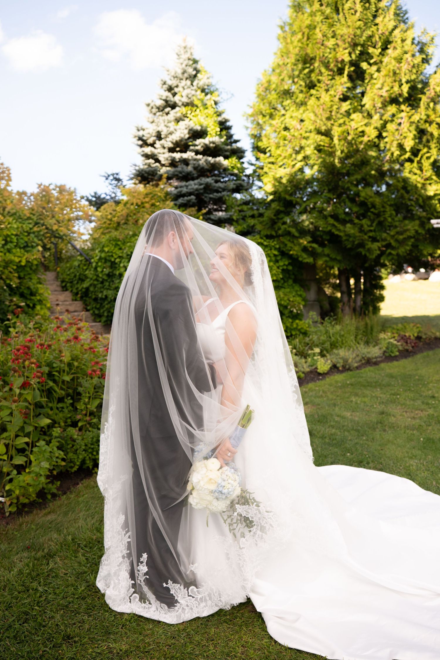 Bride and groom share a kiss under a flowing lace wedding veil in a lush garden setting.