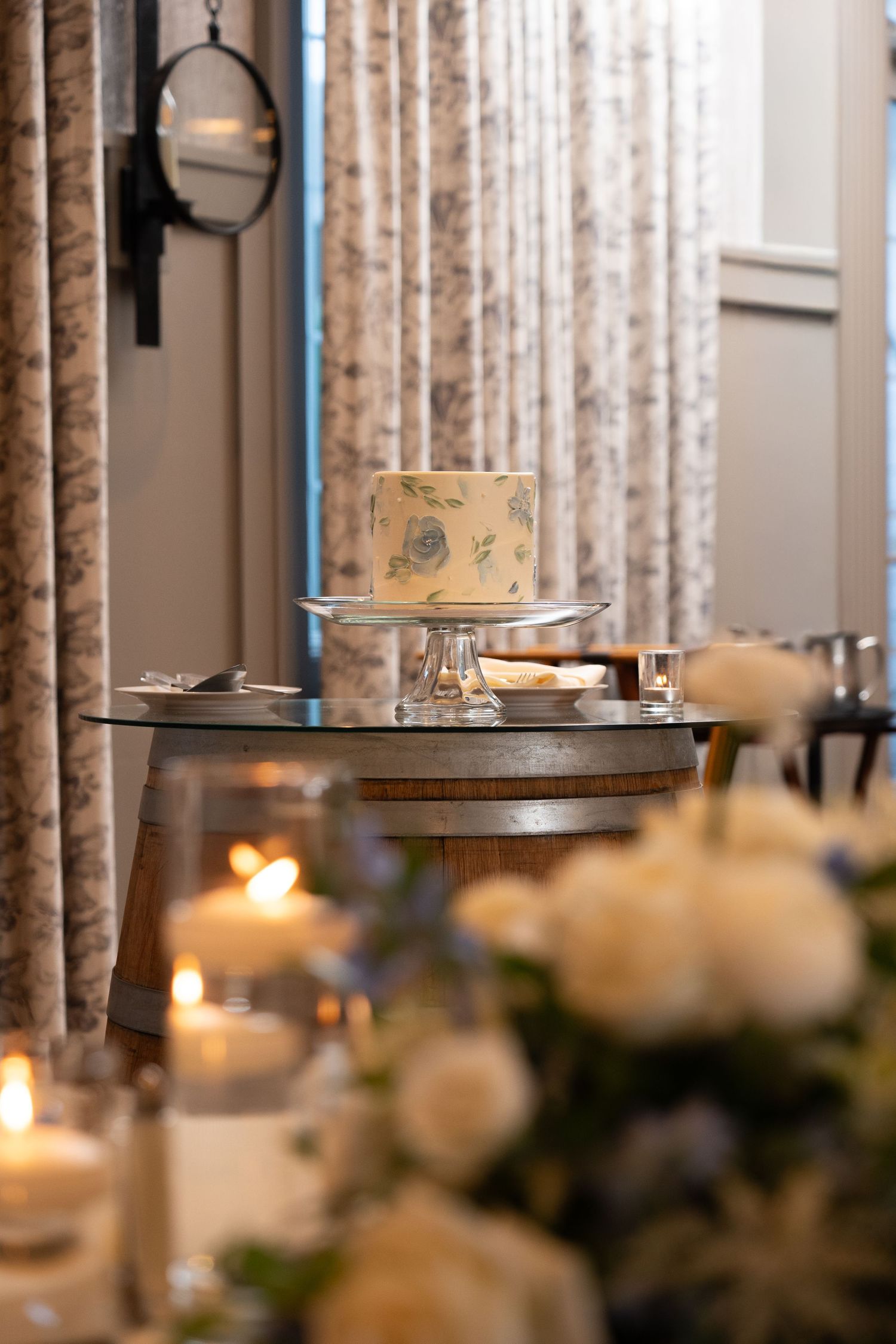 Elegant wedding cake on glass stand with lit candles and white floral arrangements in a softly lit reception venue.