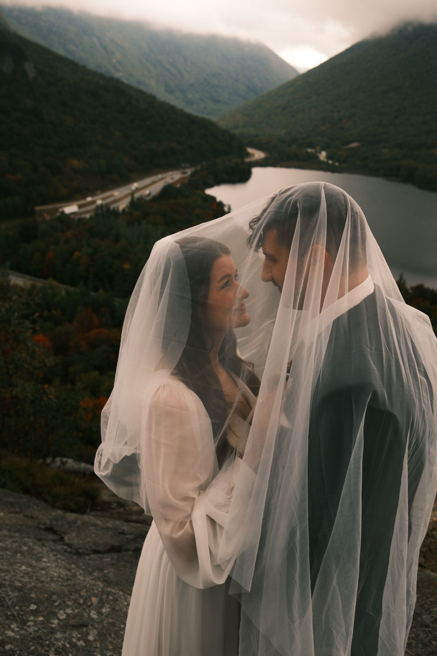 Bride and groom sharing romantic moment under wedding veil with scenic mountain lake view behind them.