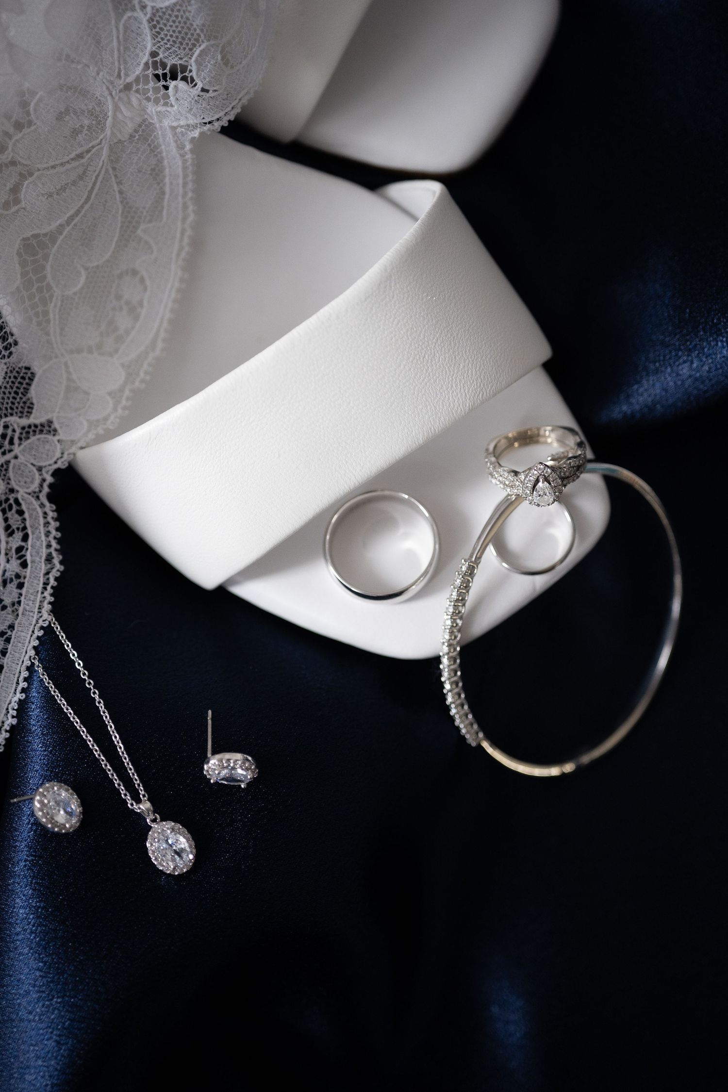Wedding jewelry flat lay featuring diamond rings, bracelet, earrings and necklace on navy satin fabric.