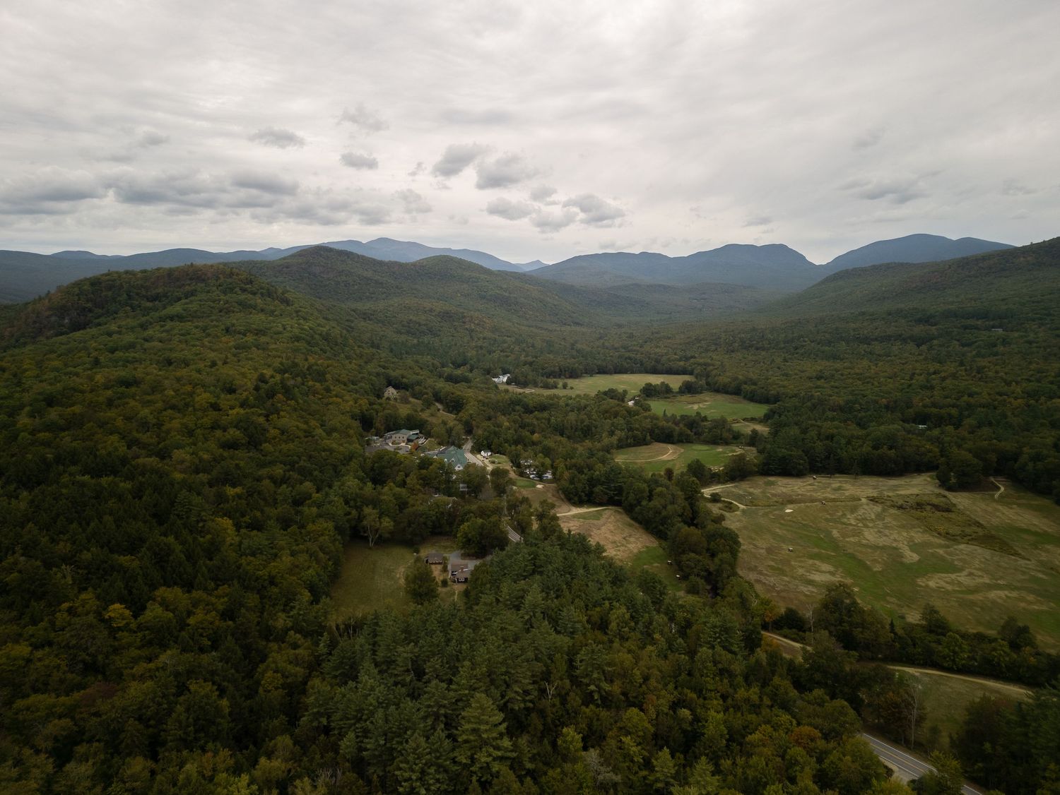 Aerial view of a lush green forested valley with scattered buildings and mountains under cloudy skies.