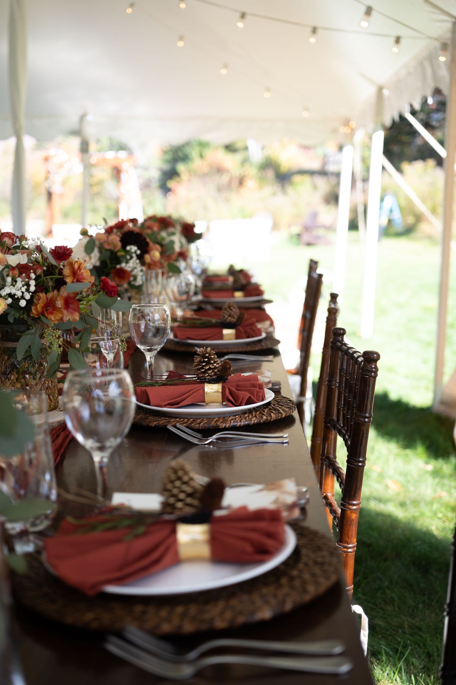 Elegant fall wedding table setting with rust napkins, pinecones, wicker chargers, and floral centerpieces under a white tent.