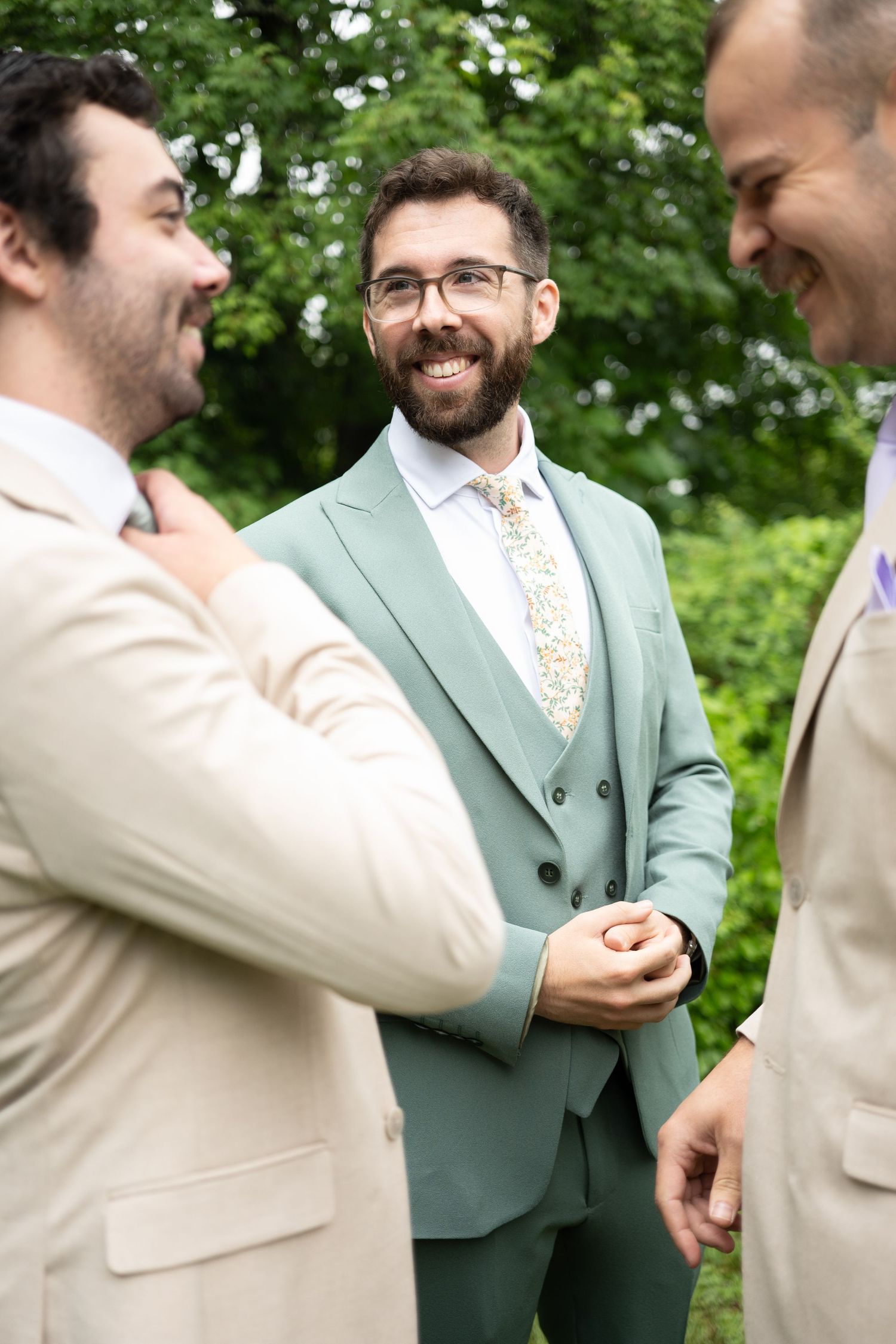 Groom in sage green suit laughs with groomsmen in tan suits outdoors at wedding.