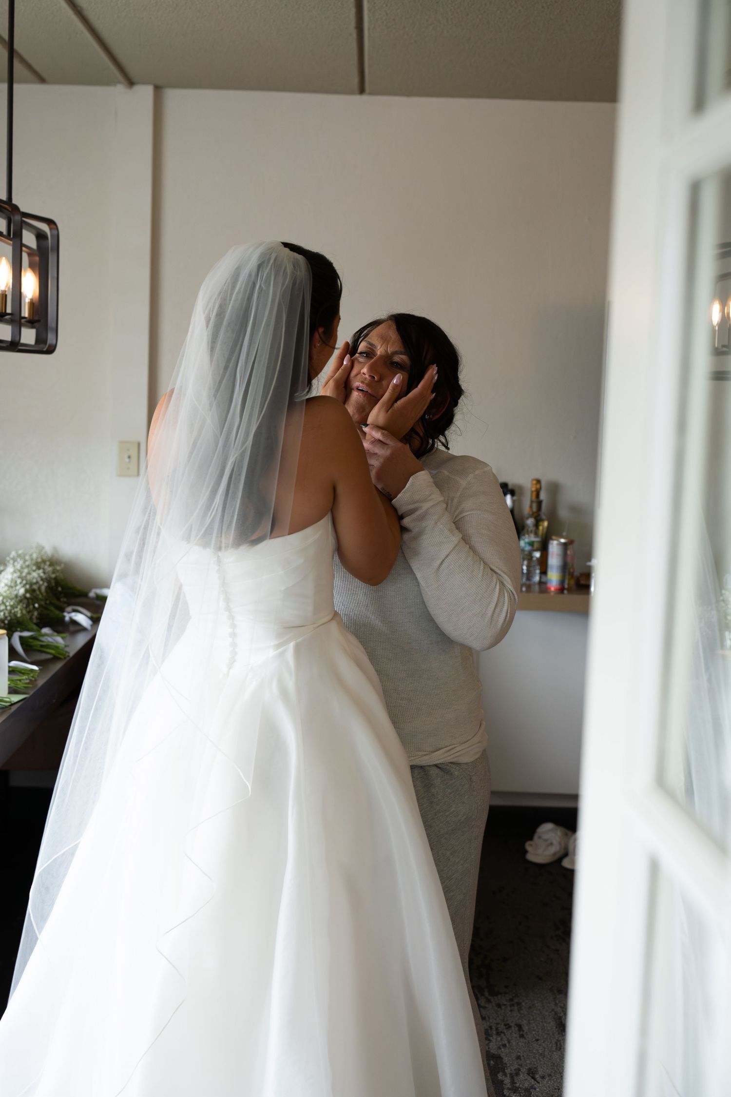 Emotional mother wiping tears while bride in white strapless gown and veil holds her face tenderly.