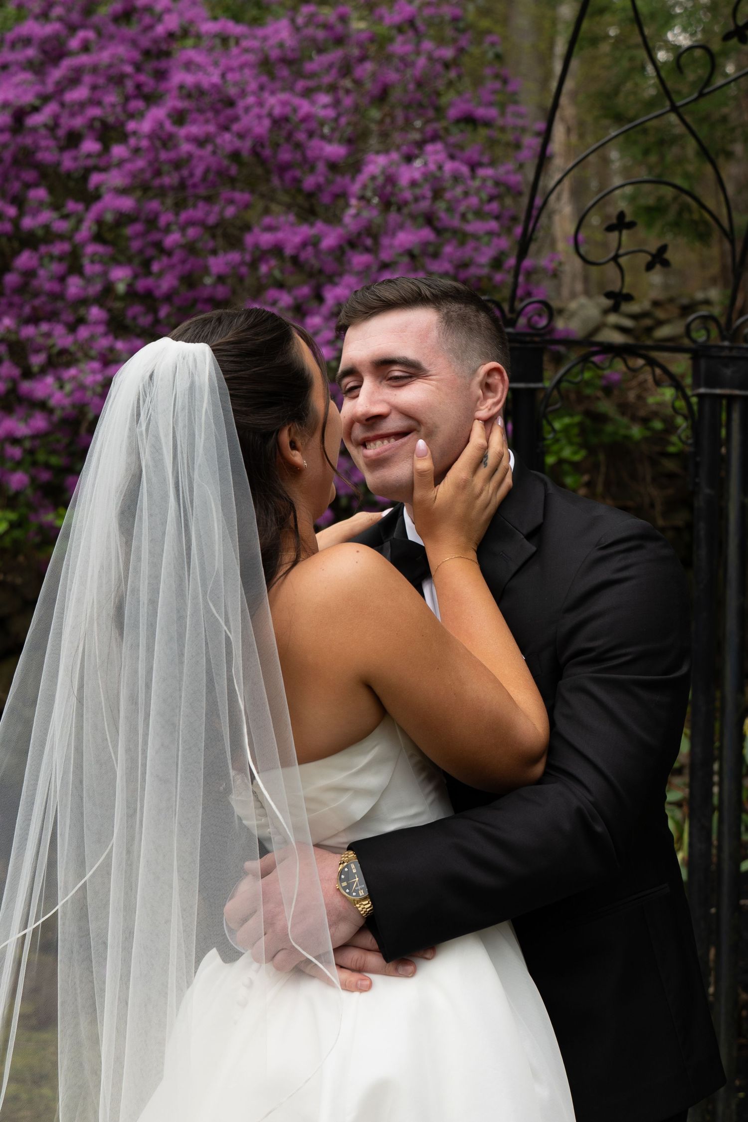 Smiling groom embraces bride in white veil before blooming purple azalea bushes in romantic outdoor portrait.