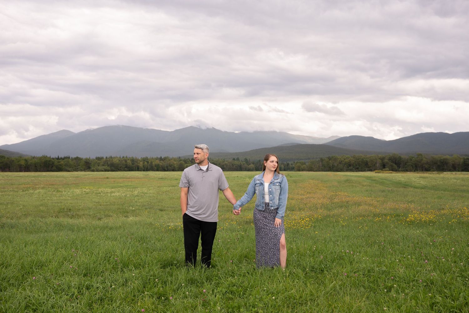 Couple holding hands in green meadow with mountain backdrop under cloudy sky.