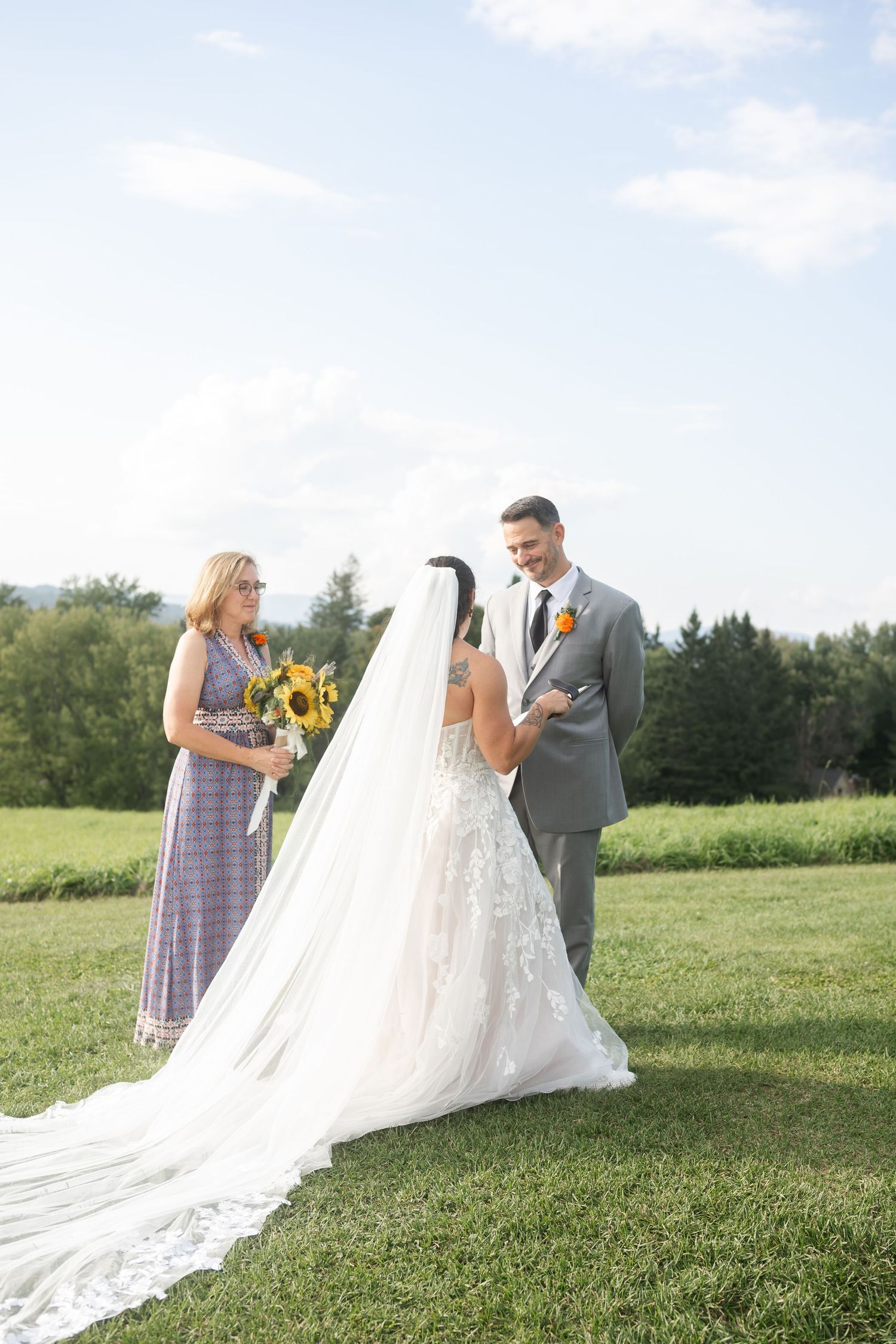 Bride and groom share a moment with bridesmaid holding sunflowers at outdoor wedding.