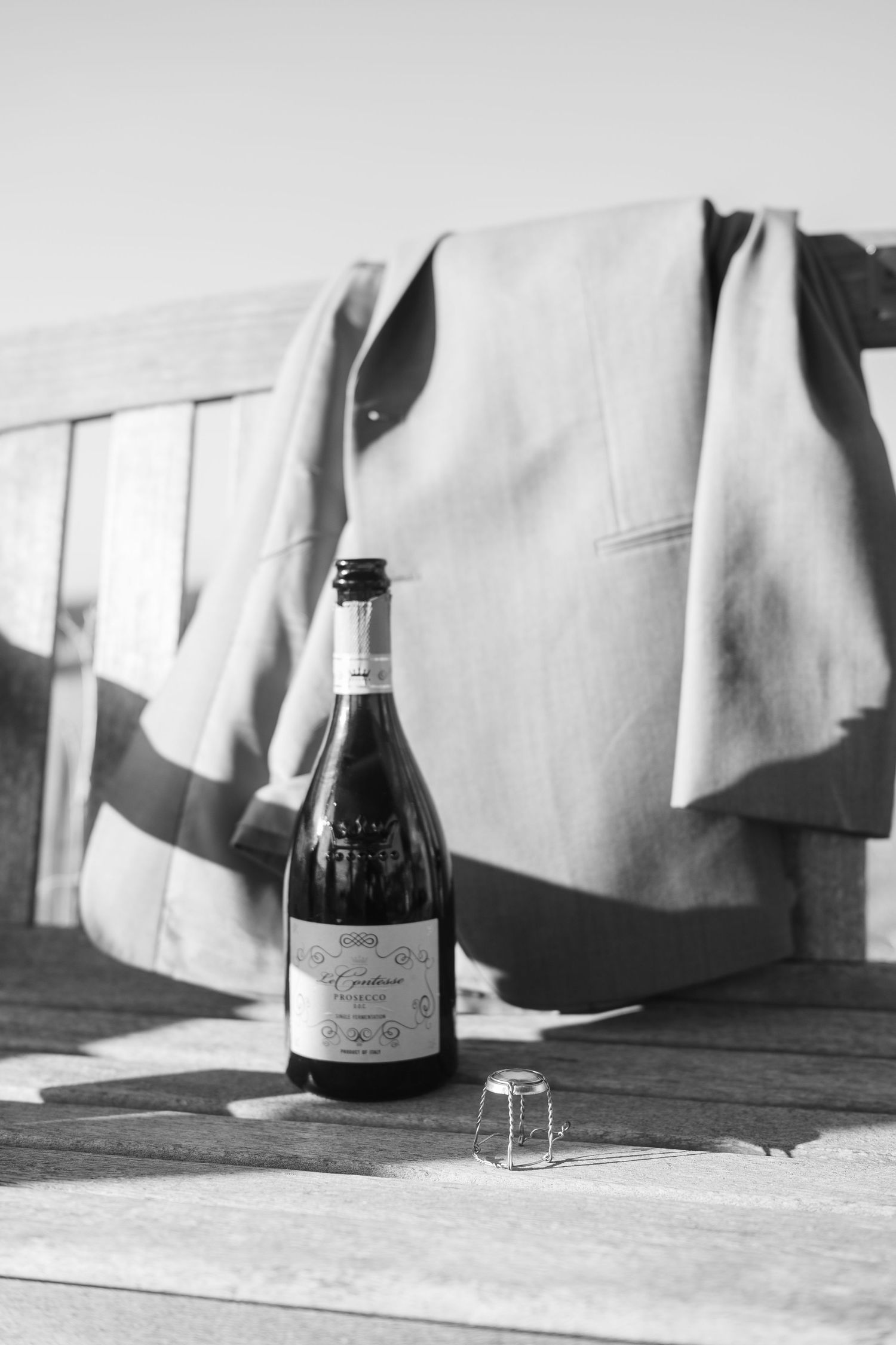 Black and white photo of a champagne bottle and cork beside a suit jacket on a bench.