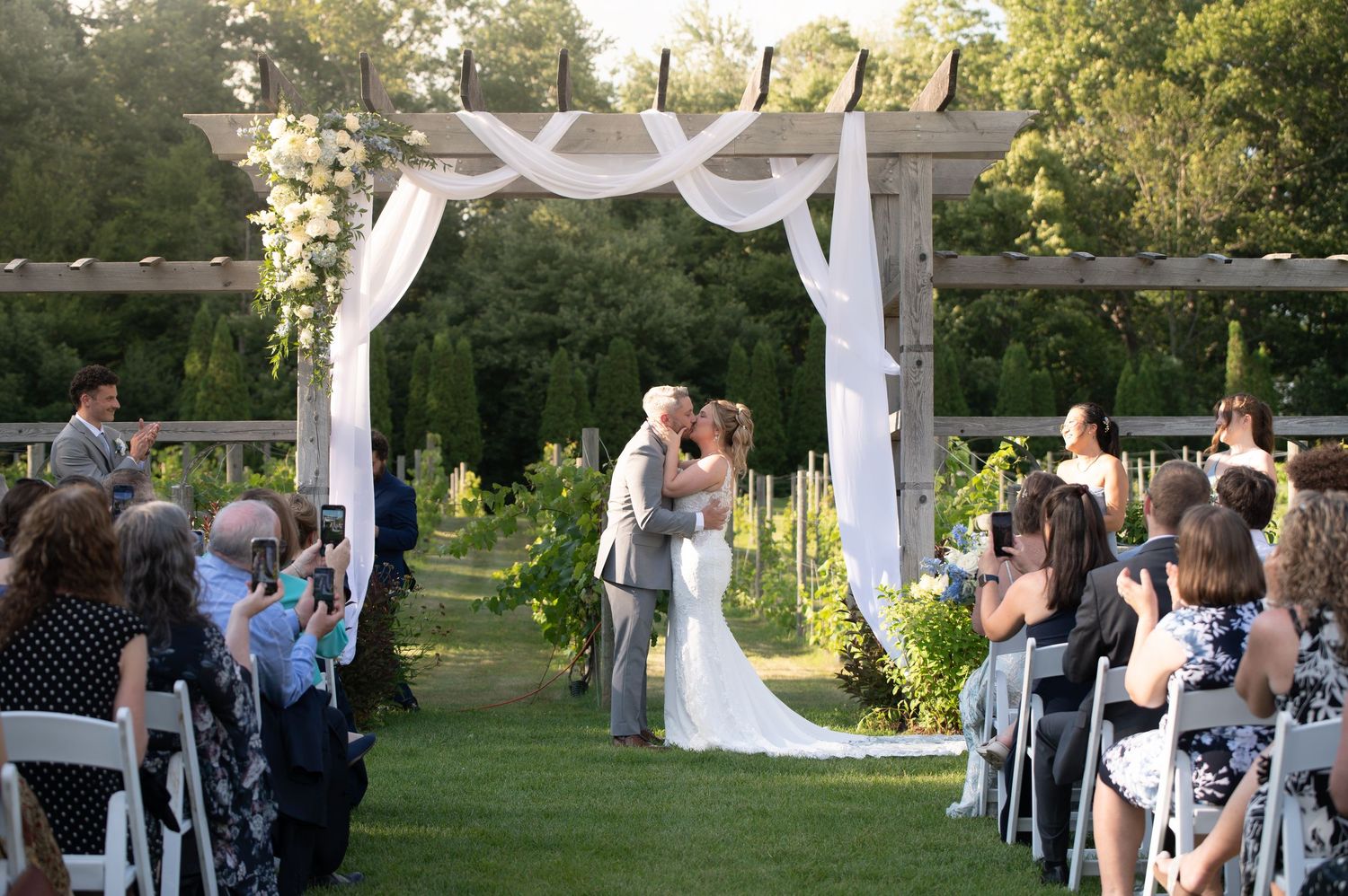 Couple shares first kiss at outdoor vineyard wedding ceremony under white draped arch with floral accents and seated guests.