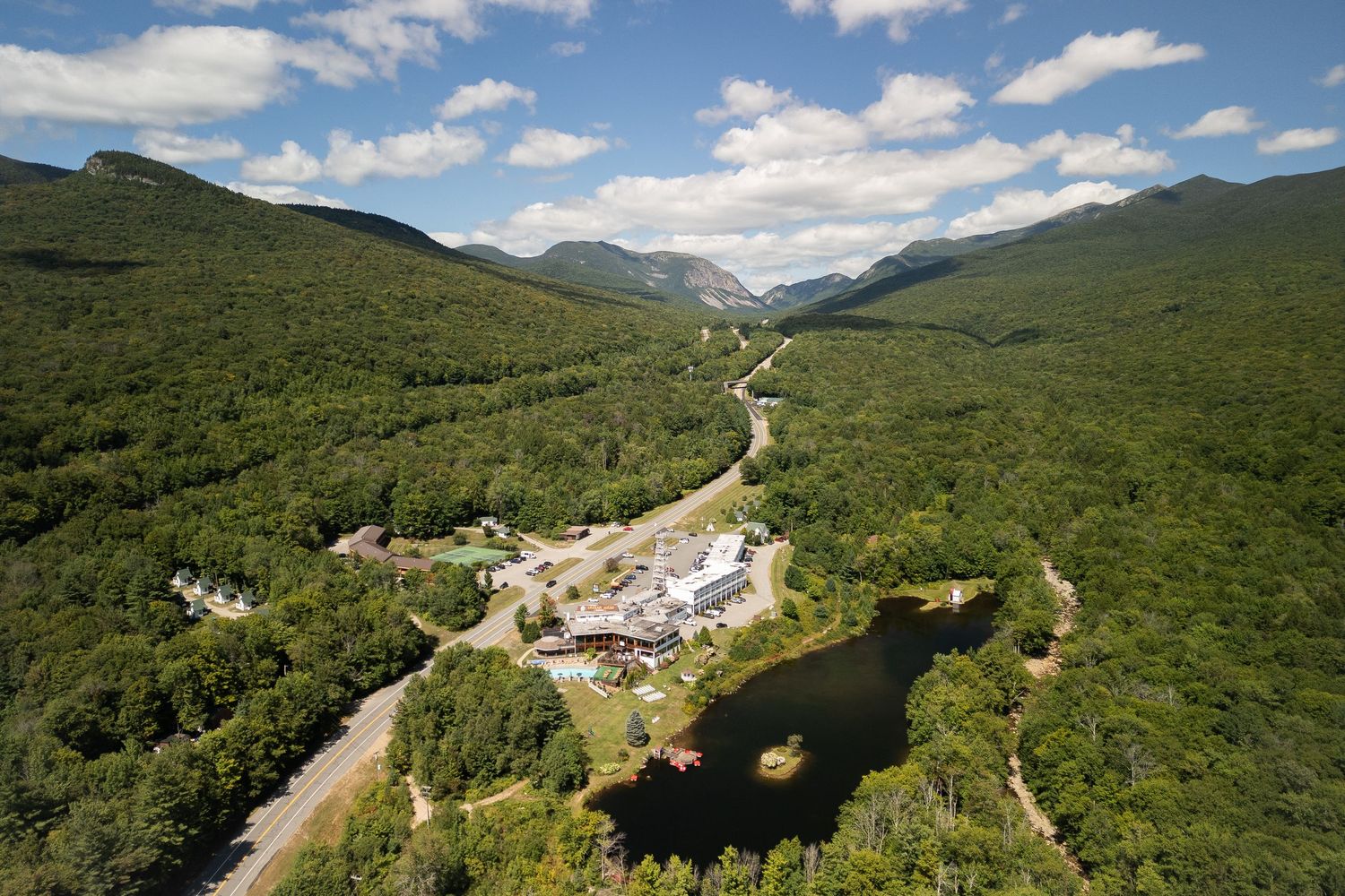 Aerial view of a mountain valley resort surrounded by lush green forests and scenic peaks under a partly cloudy sky.