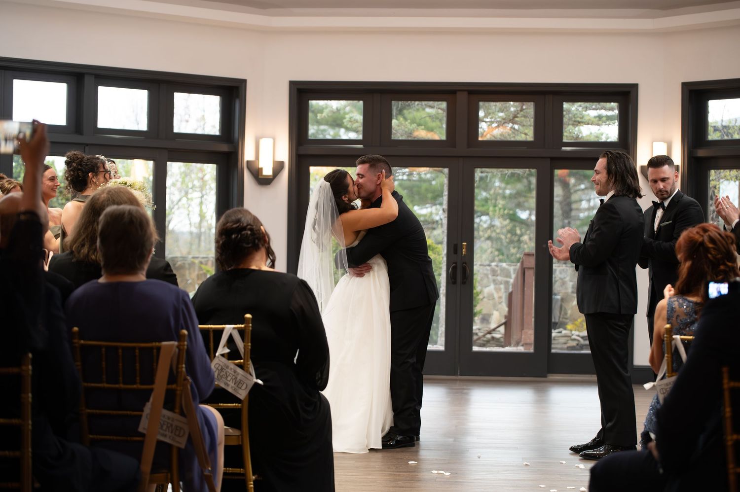 Newlyweds share first kiss as husband and wife at indoor wedding ceremony with cheering guests.