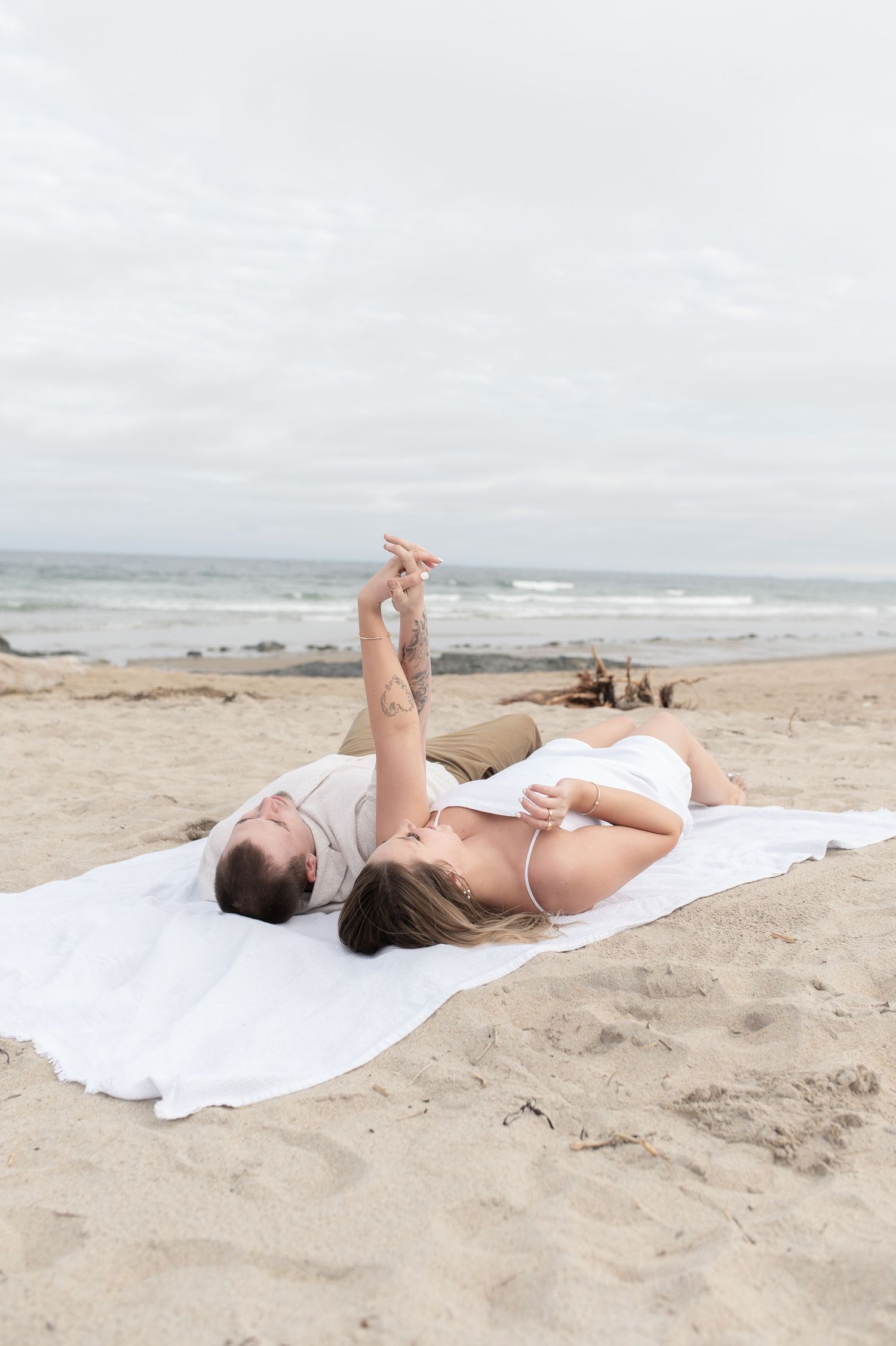 A couple lies on a white blanket on the beach, relaxing and pointing at the sky.