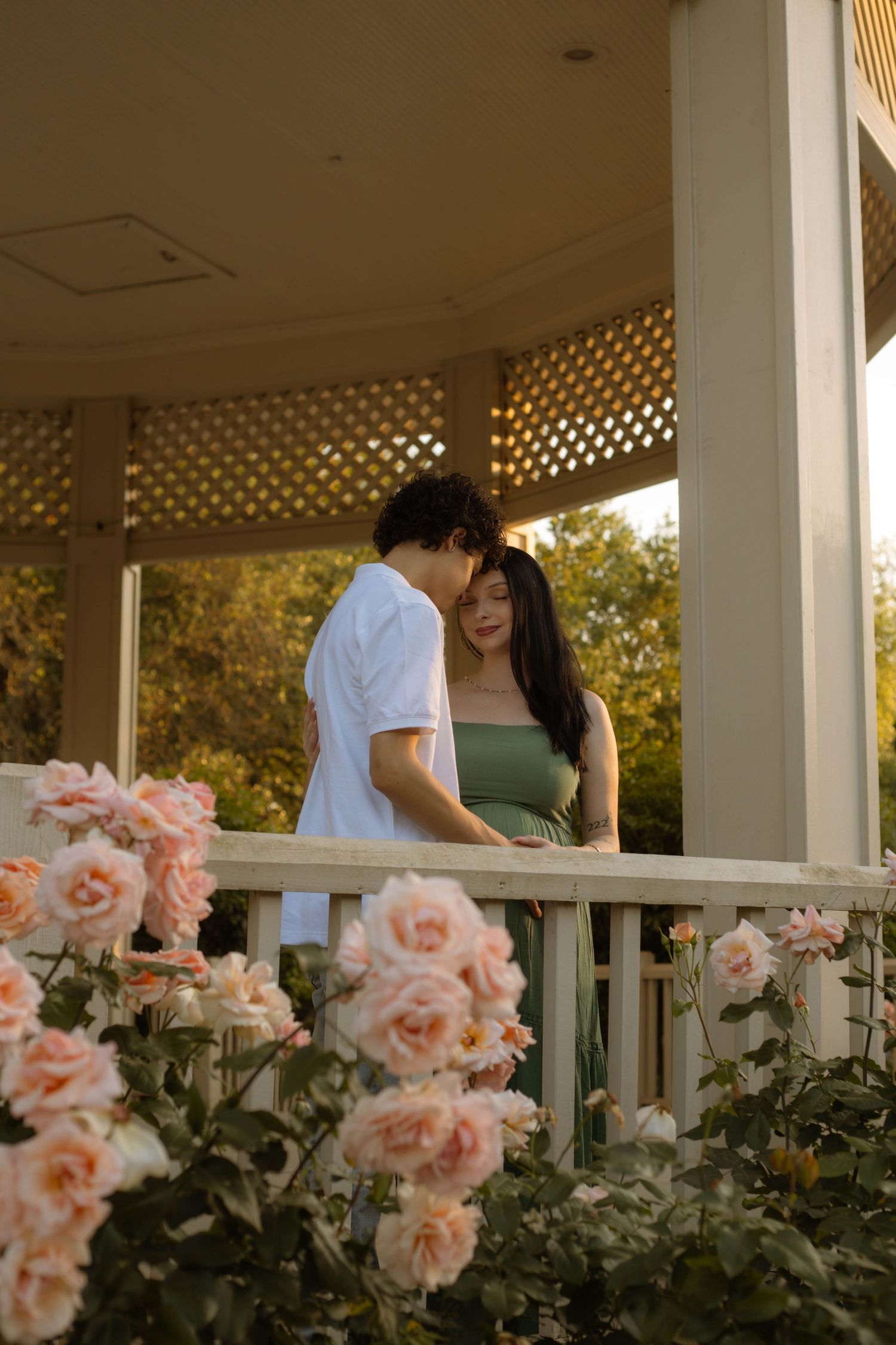 A couple shares a romantic moment on a flower-lined gazebo porch surrounded by blooming pink roses at golden hour.