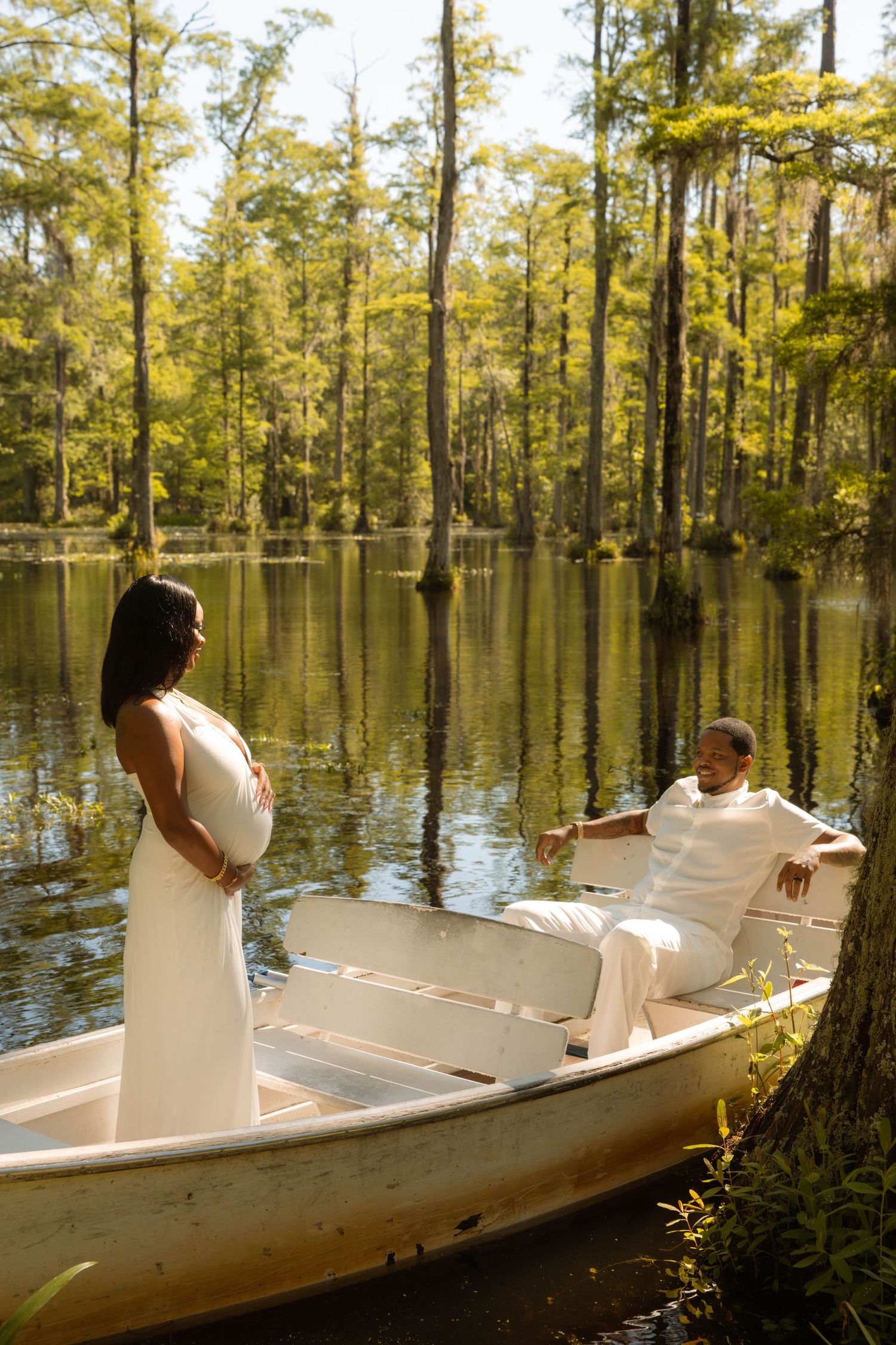 Couple in white attire poses romantically in a rowboat on a serene cypress tree-lined lake.