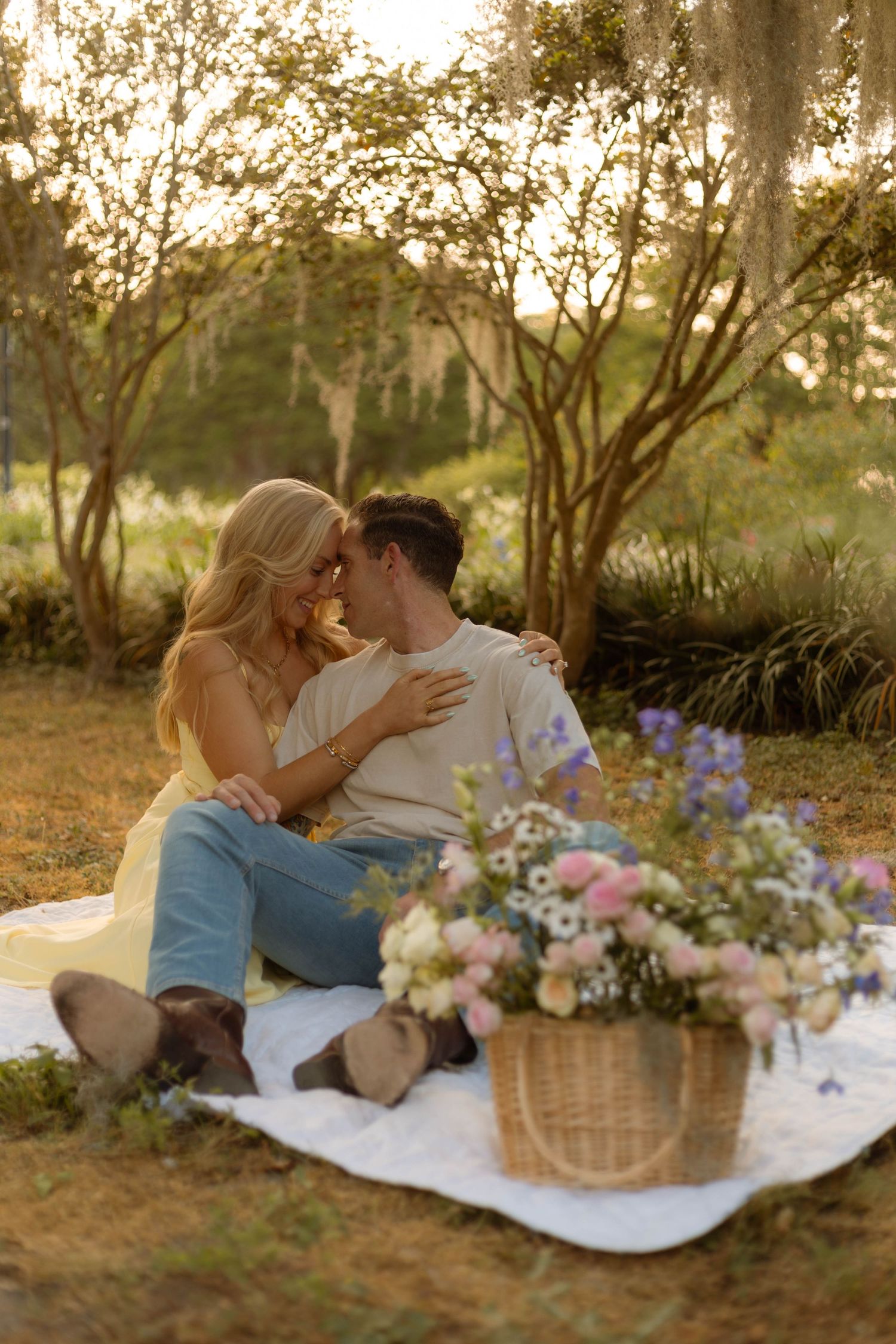 Romantic couple sits close on white blanket at golden hour with wildflower basket in lush garden setting.