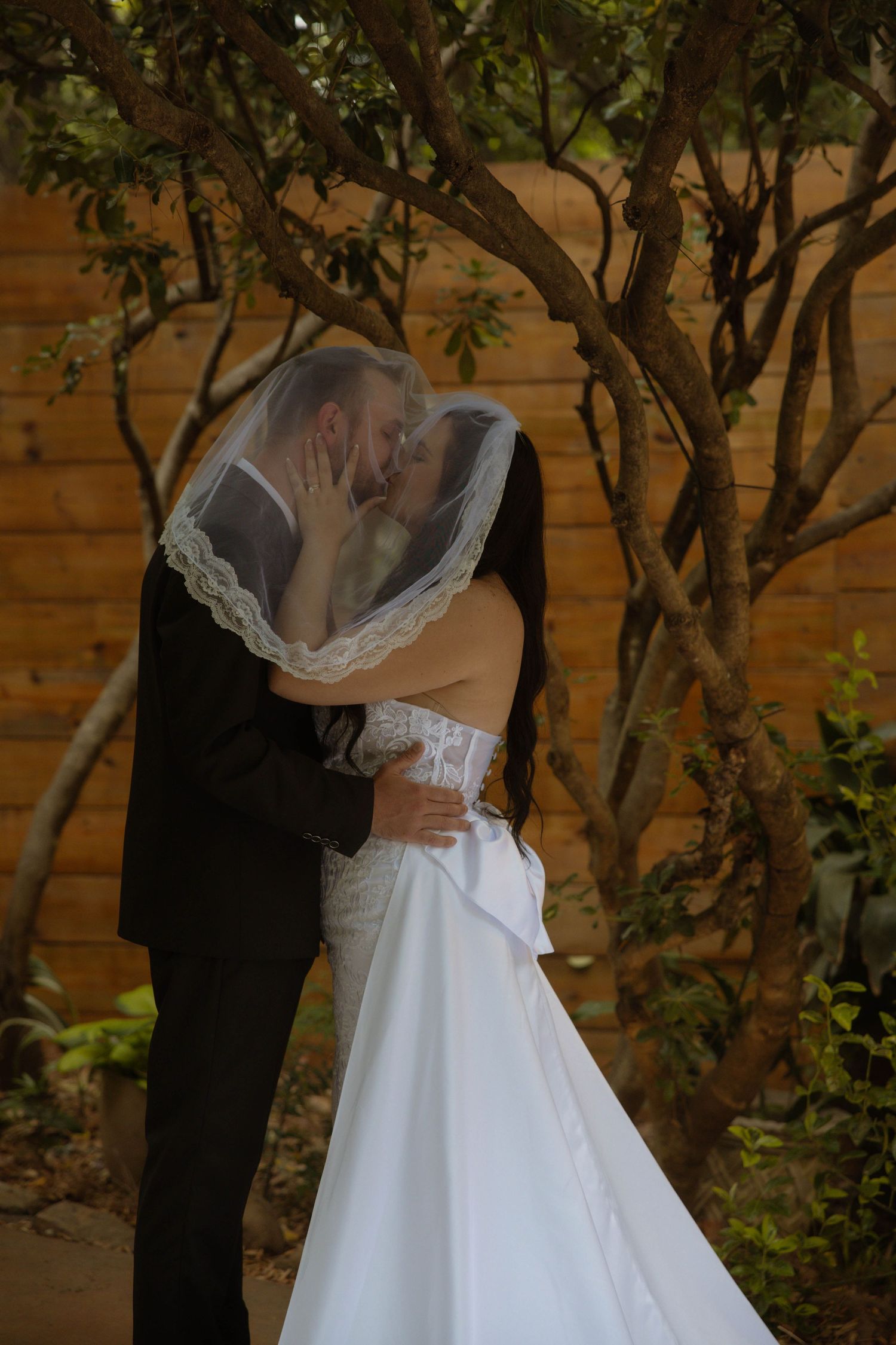 Bride and groom sharing a kiss under a wedding veil, surrounded by trees and a wooden wall backdrop.
