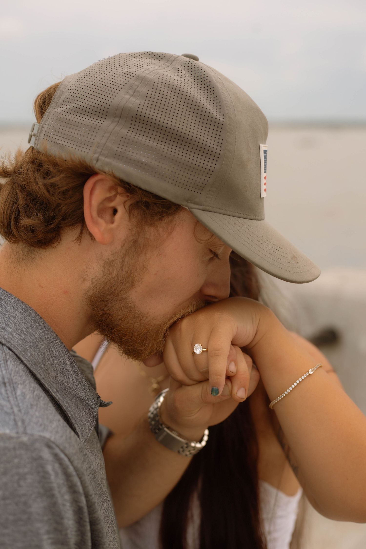 A bearded man in a gray cap tenderly kisses a woman's hand adorned with a diamond ring near the ocean.