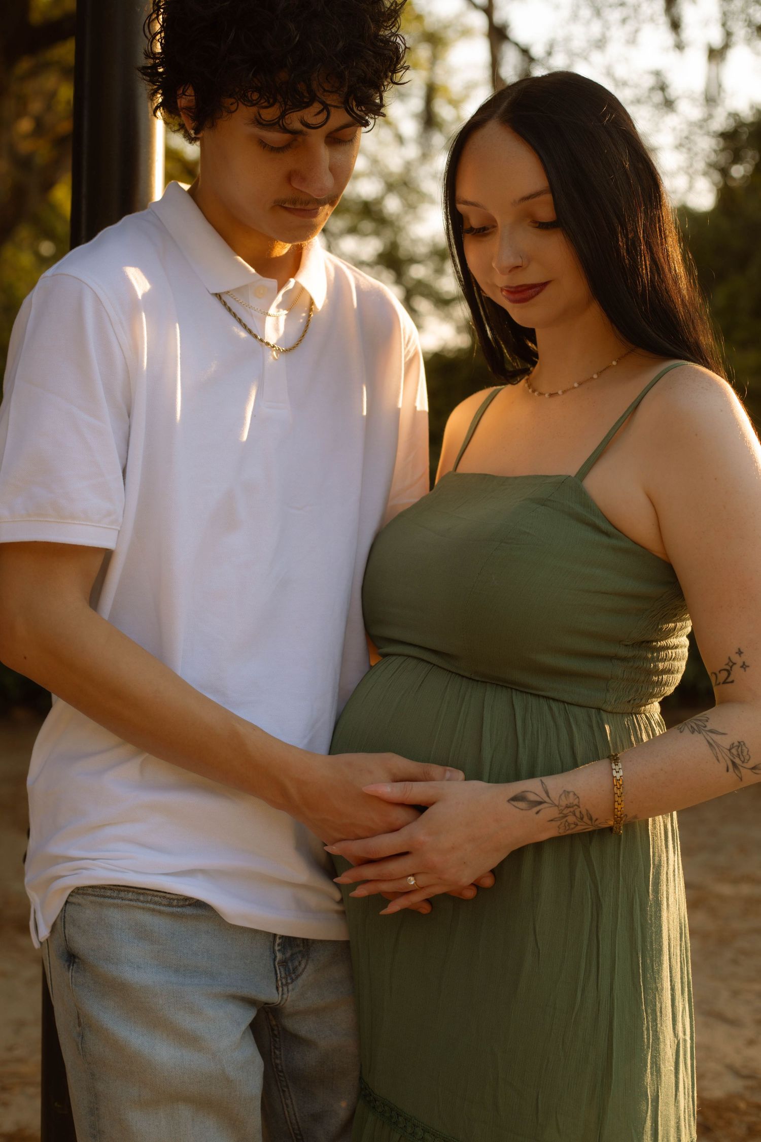 Couple tenderly holding pregnant woman's belly outdoors in golden light, man in white shirt, woman in sage green maternity dress.