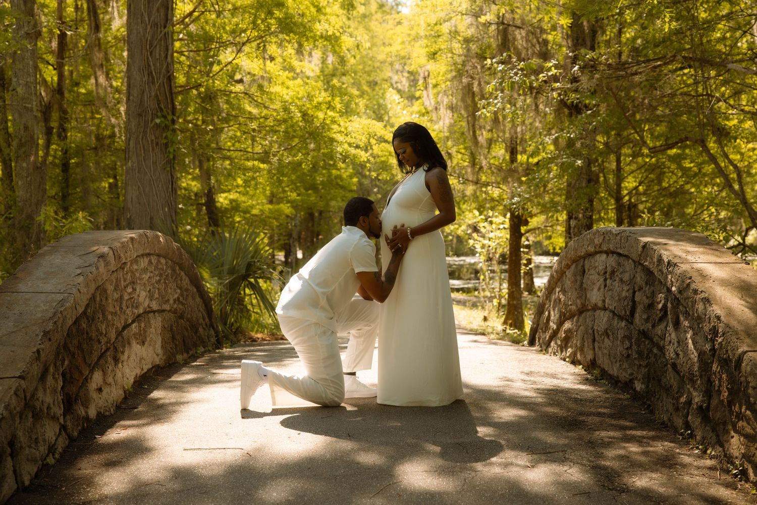 Couple in white attire on stone bridge in lush forest, man kneeling before pregnant woman.
