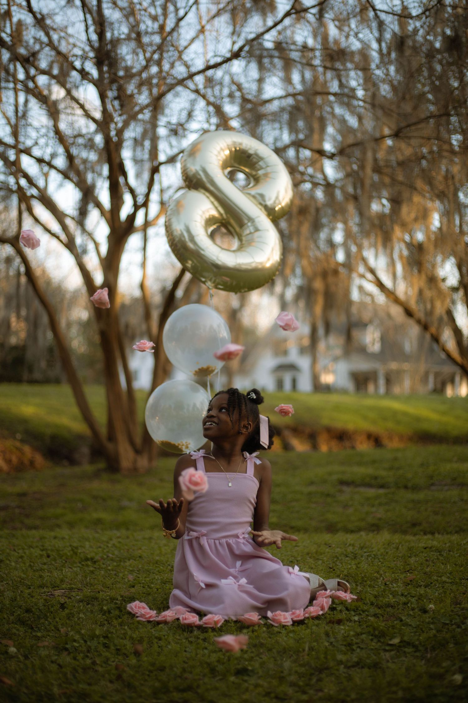 A young girl in a pink dress sits on grass outdoors, celebrating her 8th birthday with gold and white balloons.