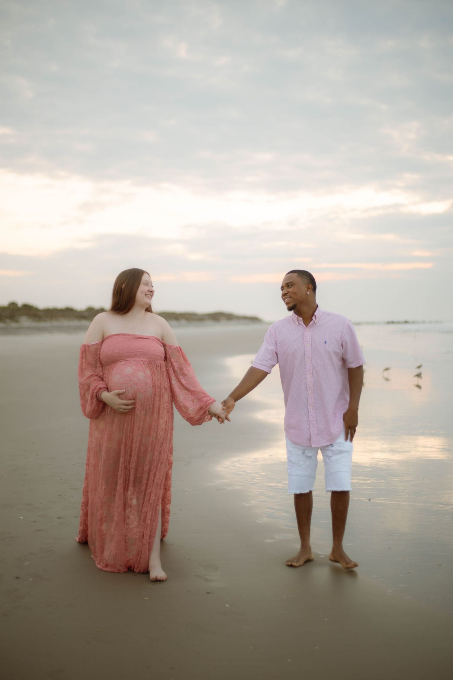Pregnant couple holding hands on beach at sunset, woman in pink maternity gown, man in pink shirt and white shorts.