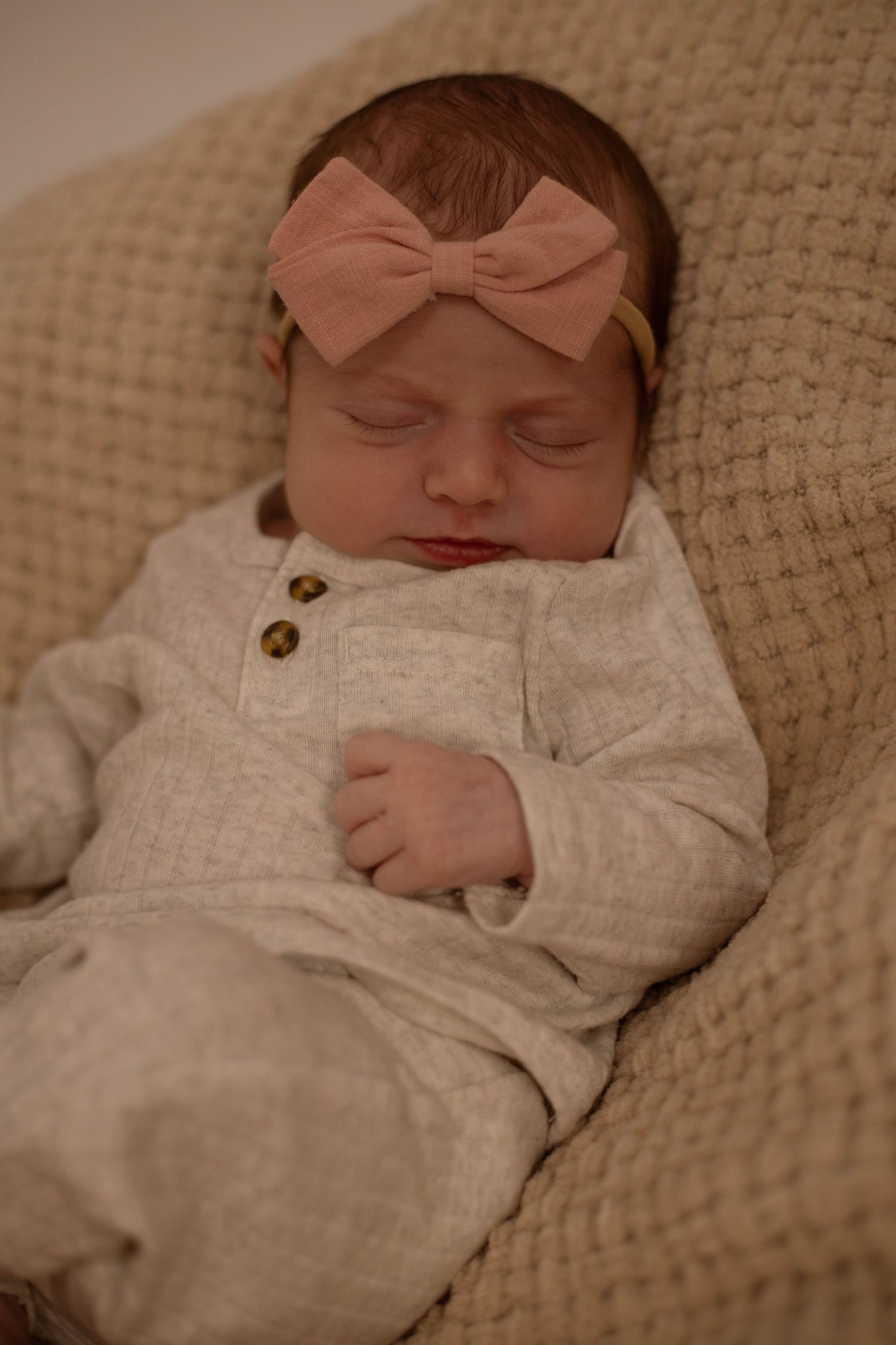 Peaceful newborn baby girl sleeping in a cream outfit with a pink bow headband.