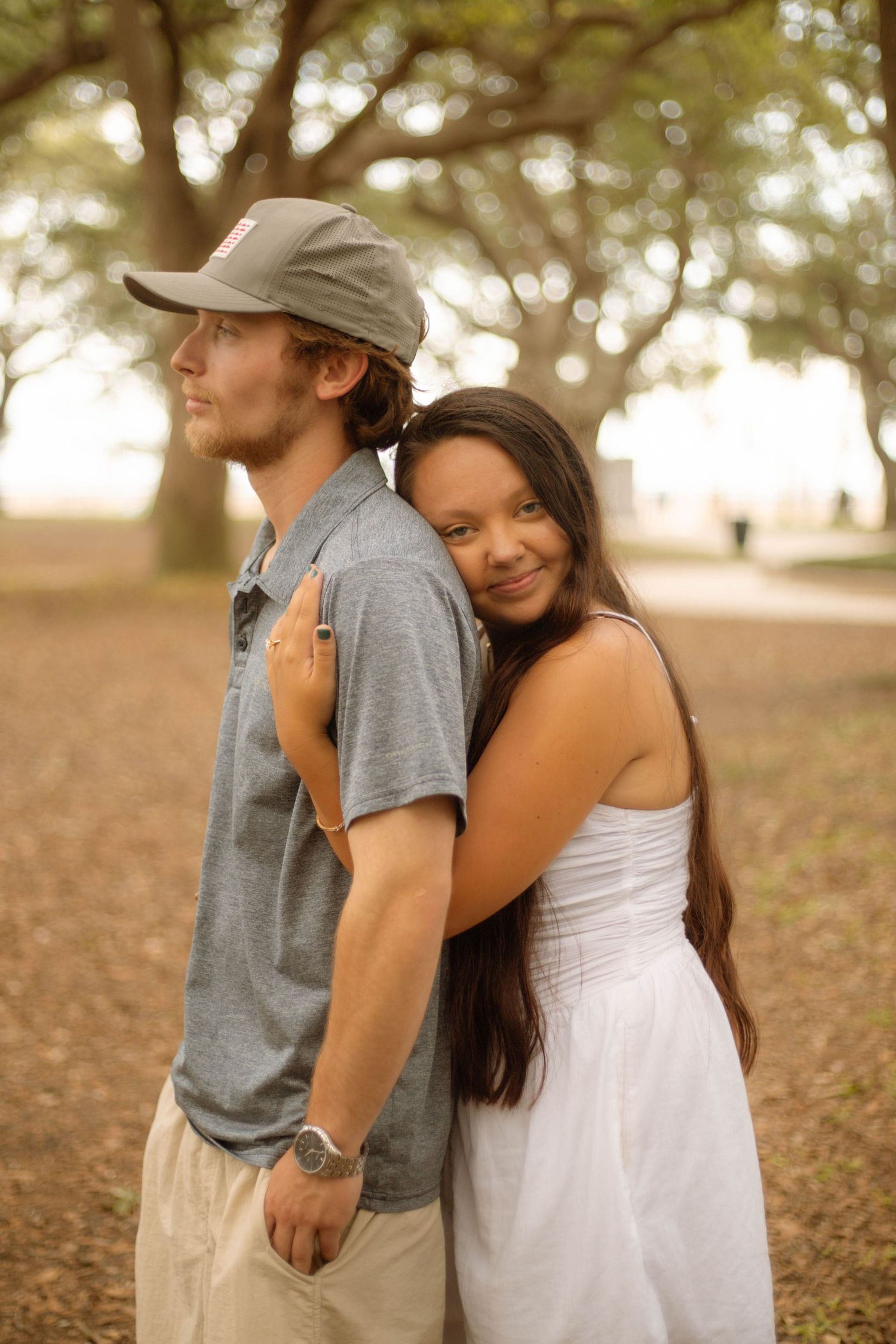 A young interracial couple poses in a park, woman in white dress smiling behind man in gray polo and cap.