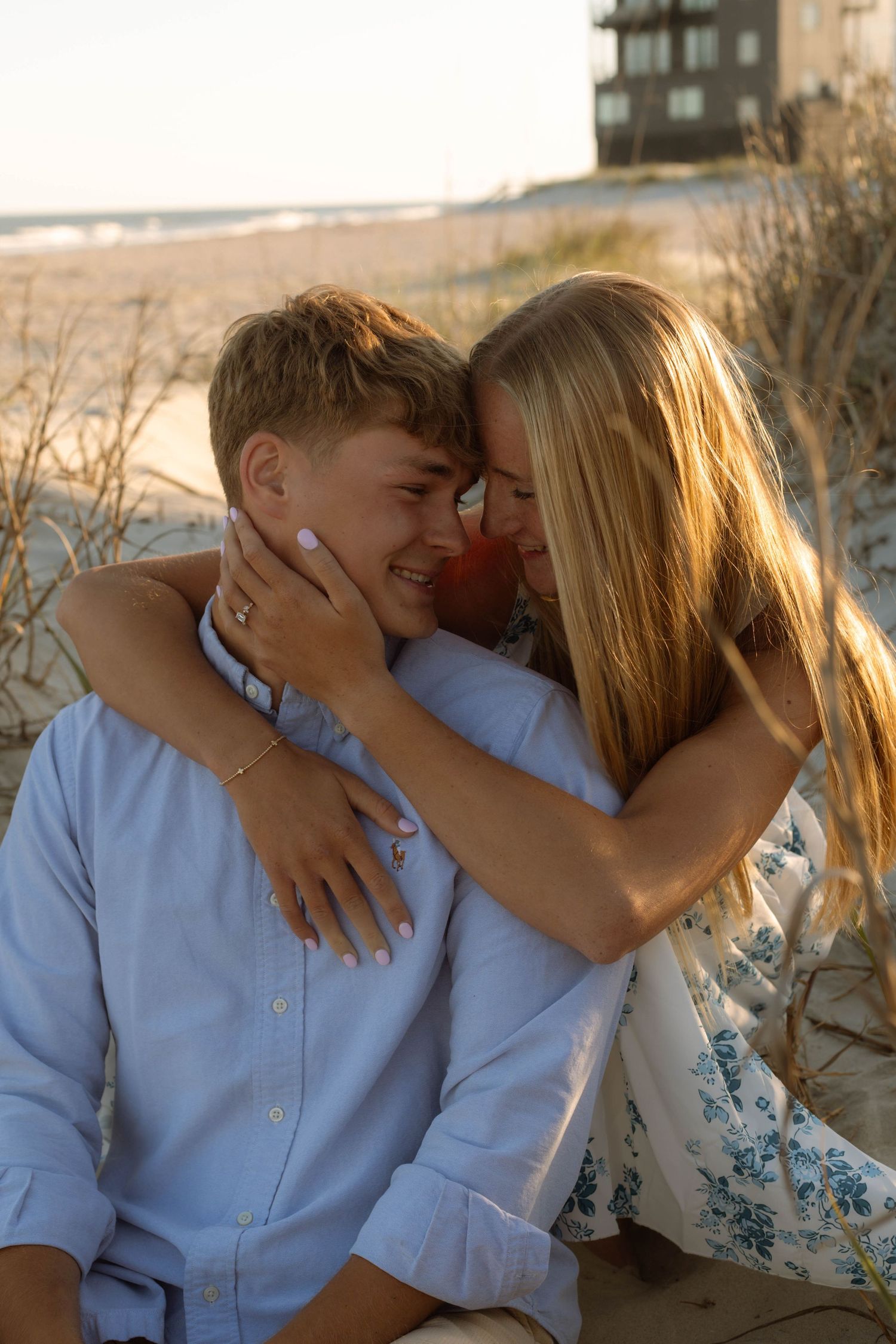Young couple sharing a romantic moment on a sandy beach at sunset, woman embracing man with foreheads touching.
