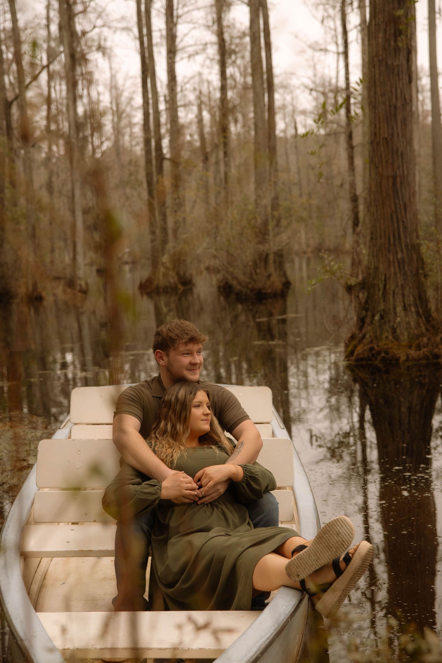Couple embracing in a white rowboat surrounded by cypress trees in a misty swamp, wearing matching olive green outfits.