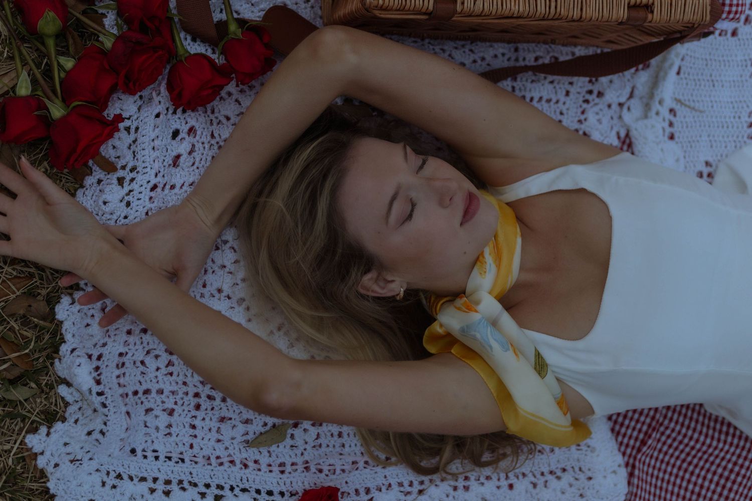 A dreamy picnic scene with red roses and a wicker basket on a white lace blanket in soft natural light.