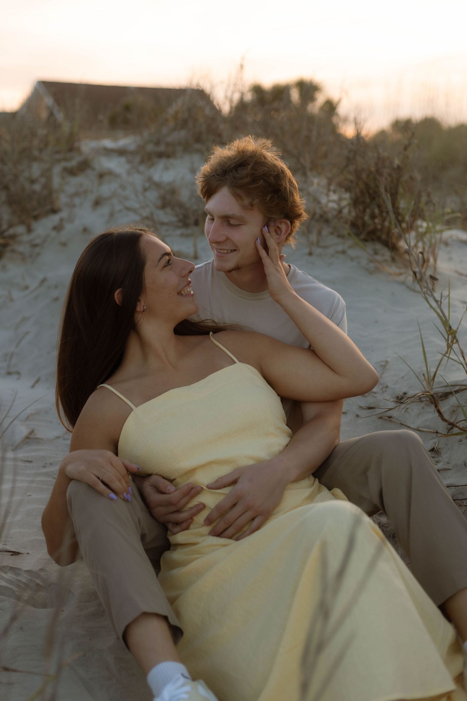 A couple sits together on sandy beach dunes at sunset, woman in yellow dress smiling as man embraces her tenderly.