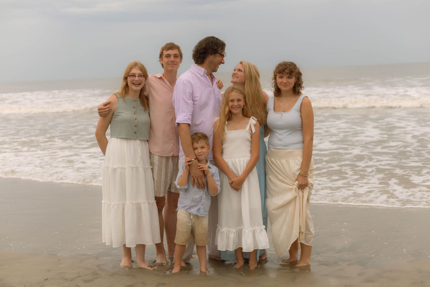 Family of seven posing together on a beach with ocean waves, wearing casual light-colored outfits for a coastal photo session.