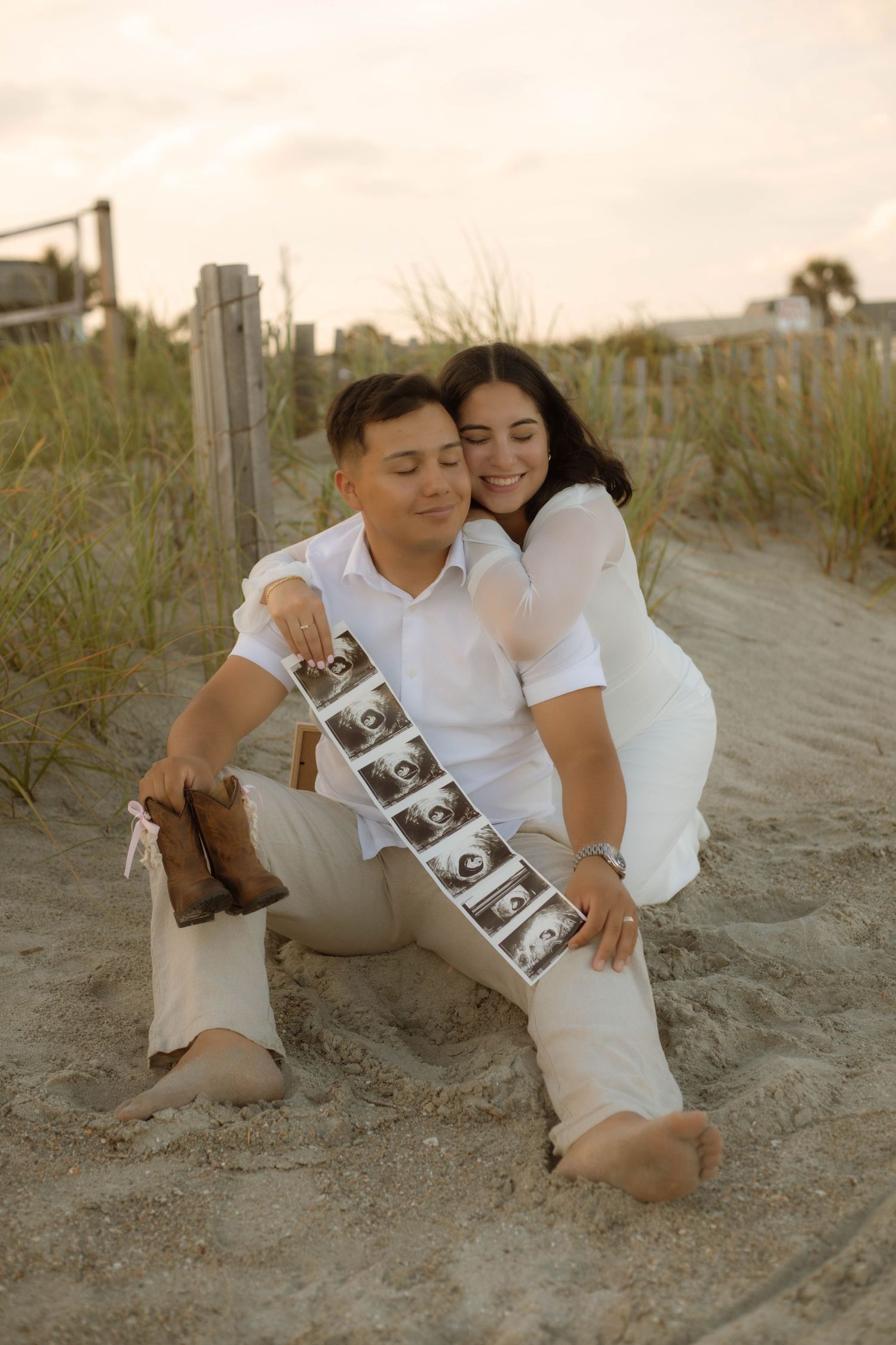 Expecting couple in white sit on beach sand, smiling while holding ultrasound photos at sunset.