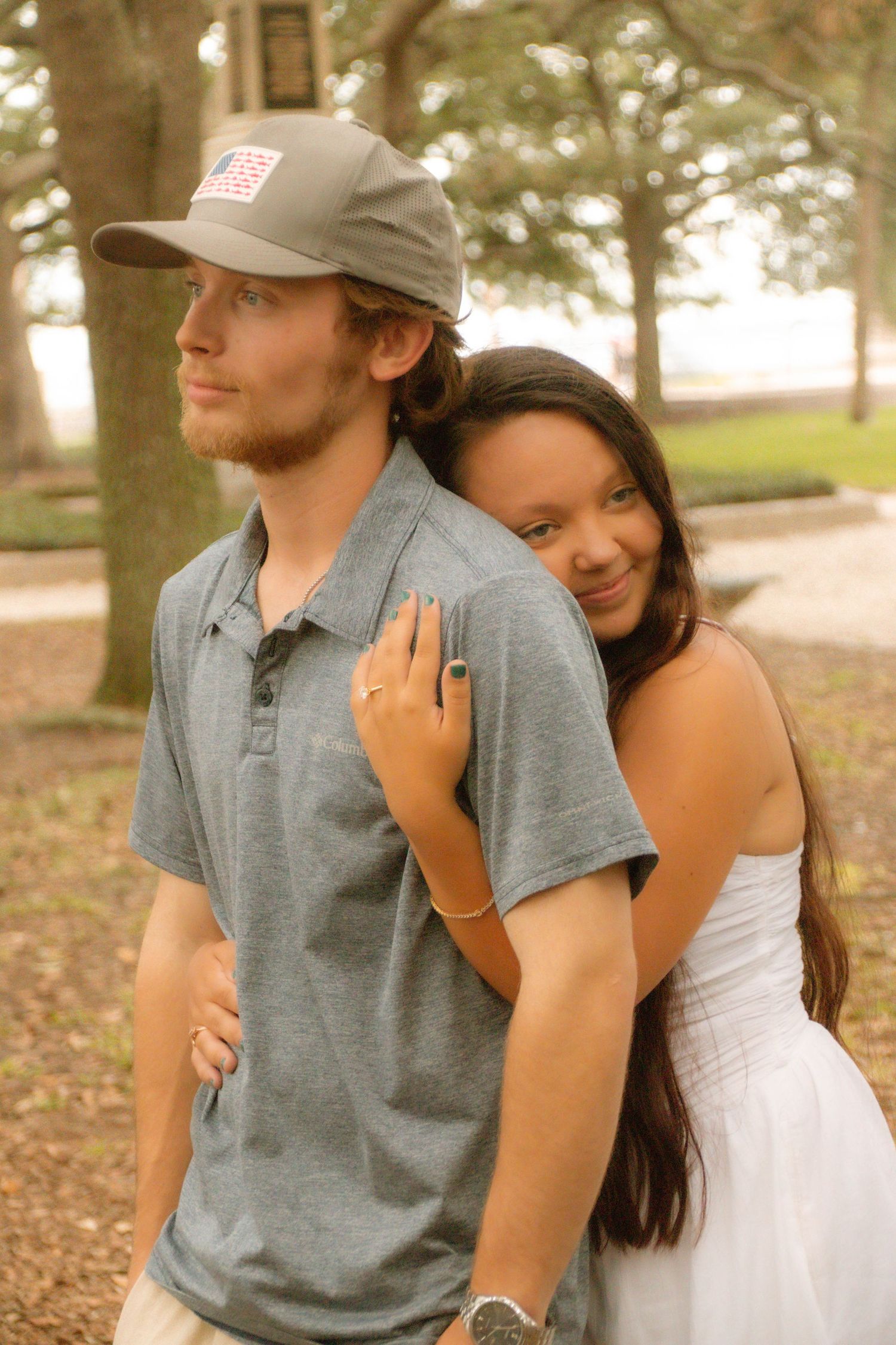 A young couple poses outdoors, woman in white dress hugging man in gray polo and cap near trees.
