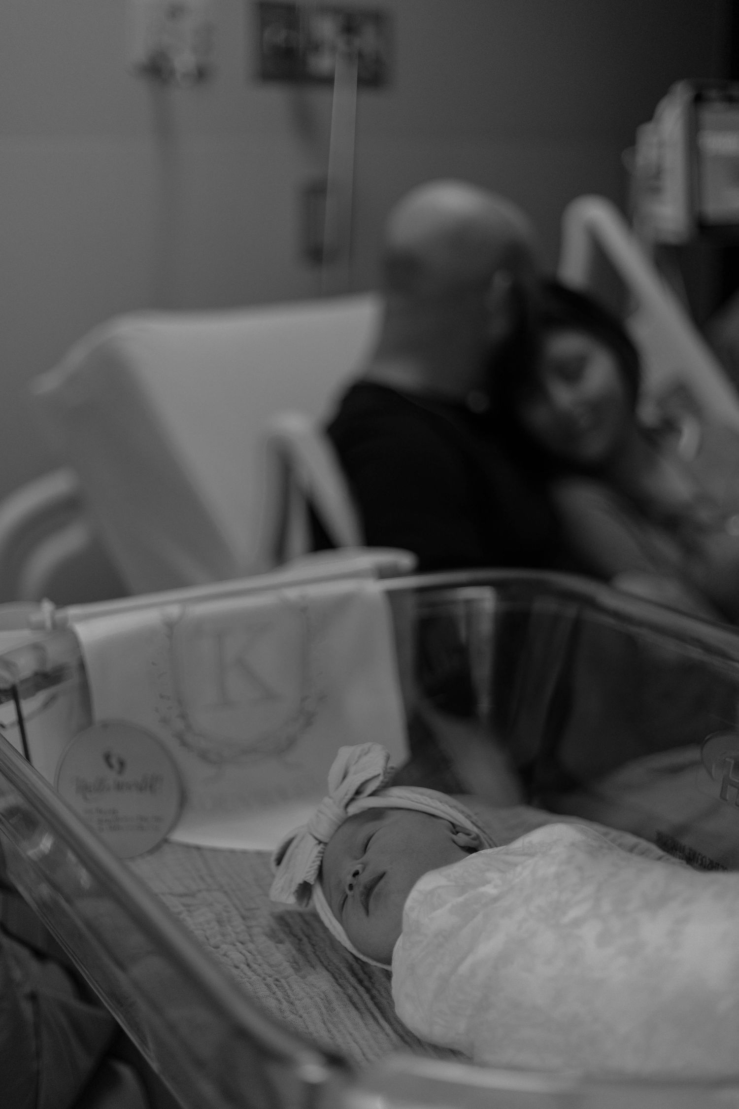 Black and white photo of a newborn baby in hospital bassinet with parents resting in background.